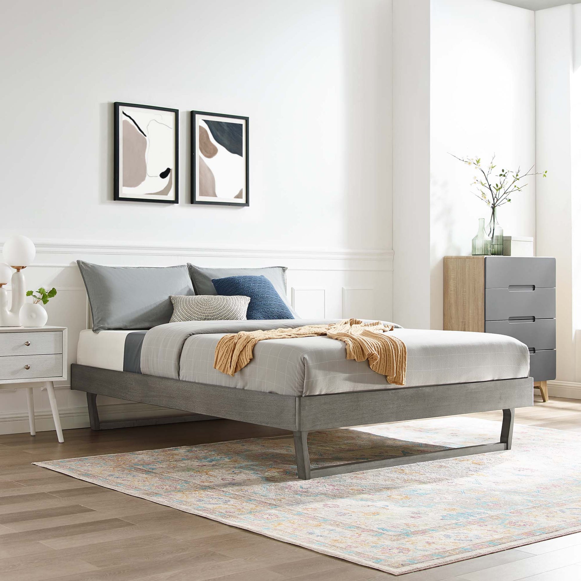 Gray wood full platform bed frame with low legs, blue-gray bedding, mustard throw, white nightstand, abstract art, pastel rug