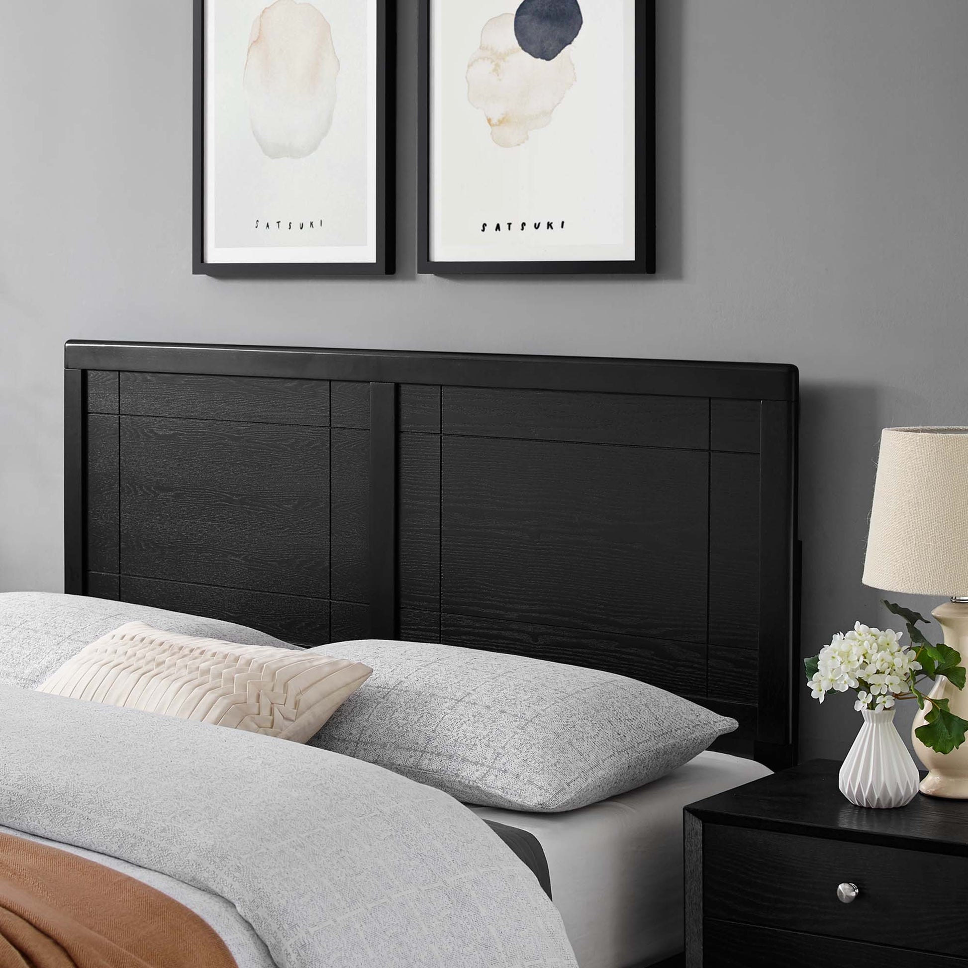 Black wood headboard with panel details, gray bedding, beige pillow, black nightstand with lamp and vase of white flowers