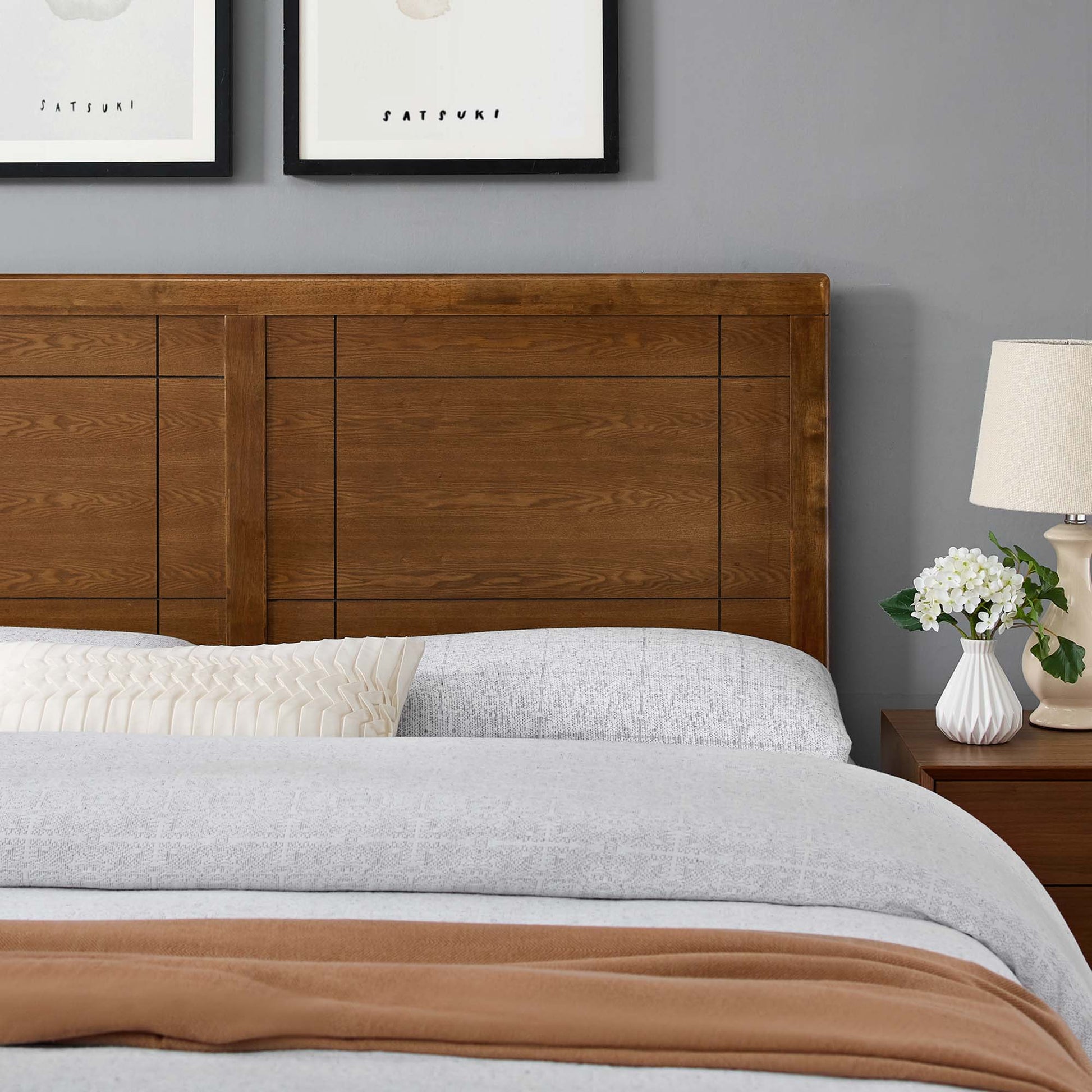 Brown wood King headboard with geometric panels, grey bedding, cream pleated pillow, rust throw; lamp and vase of white flowers.