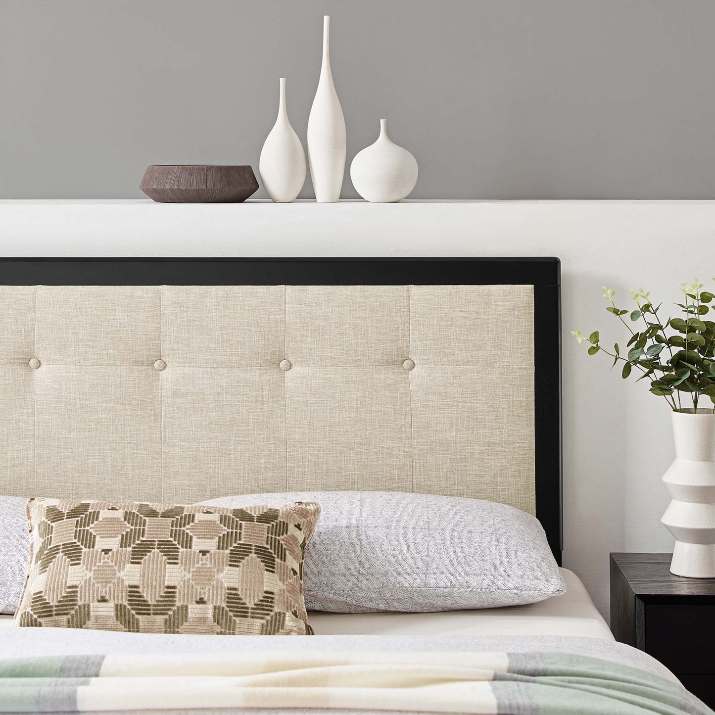 Beige tufted headboard with black frame, beige bedding and geometric pillow; white vases, dark bowl on shelf.