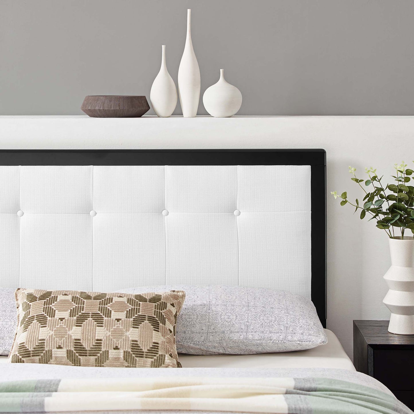 White tufted headboard with black frame, beige geometric pillow, gray bedding, white vases and plant on bedside table.