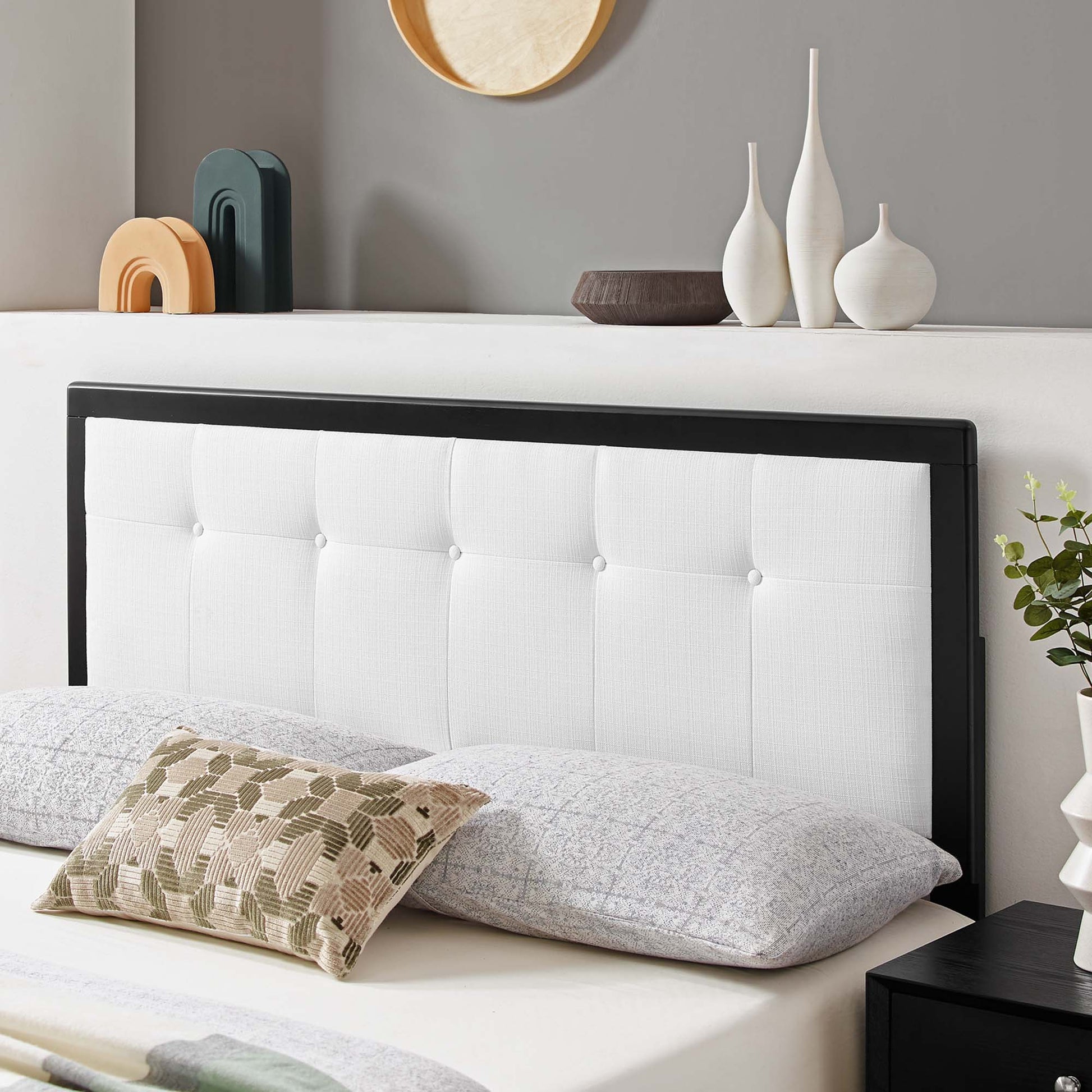 White tufted headboard with wood frame, gray bedding and a beige geometric pillow; decorative vases on a shelf.