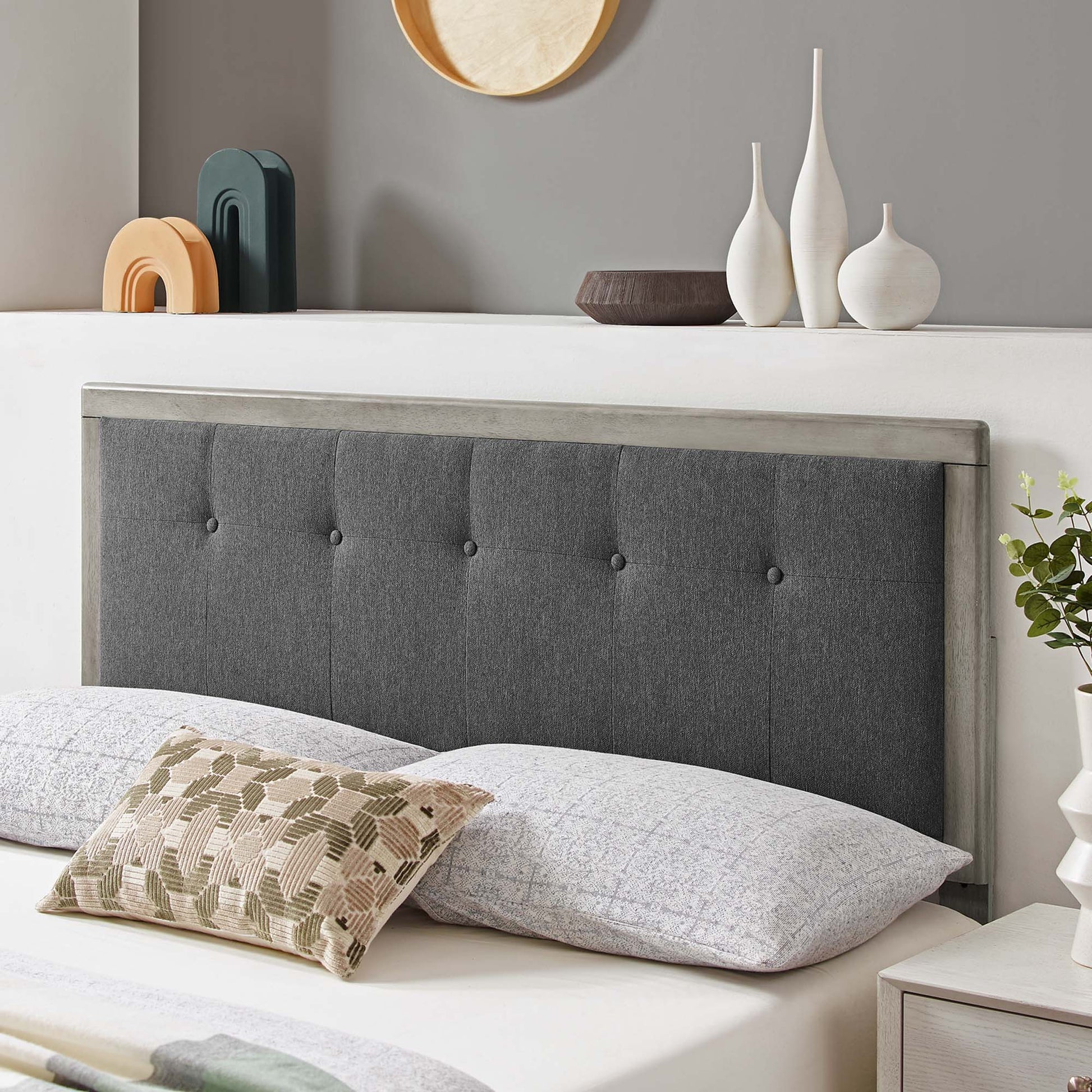 Gray tufted headboard with a wood frame, light gray bedding and a beige geometric pillow; ceramic vases and bowls on a shelf.