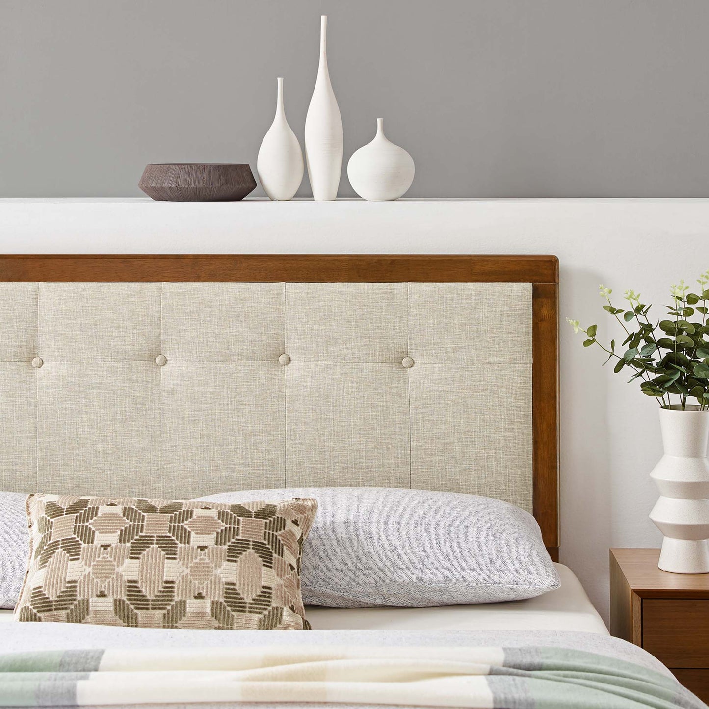 Brown-framed beige tufted king headboard with a geometric pillow, pastel throw, and white ceramic vases on a shelf beside the bed.