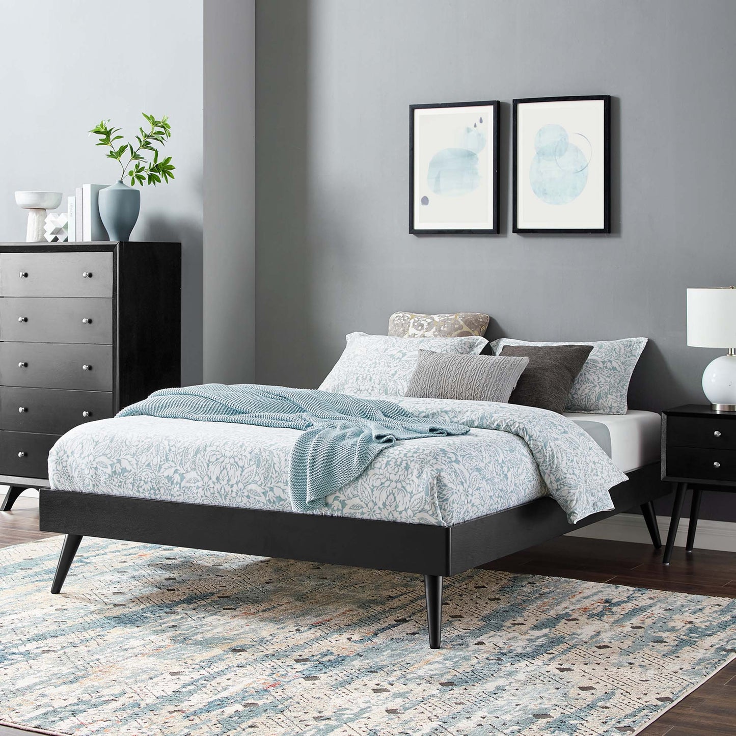 Low-profile black platform bed with tapered legs, blue floral bedding, pillows and a light blue throw in a modern bedroom.