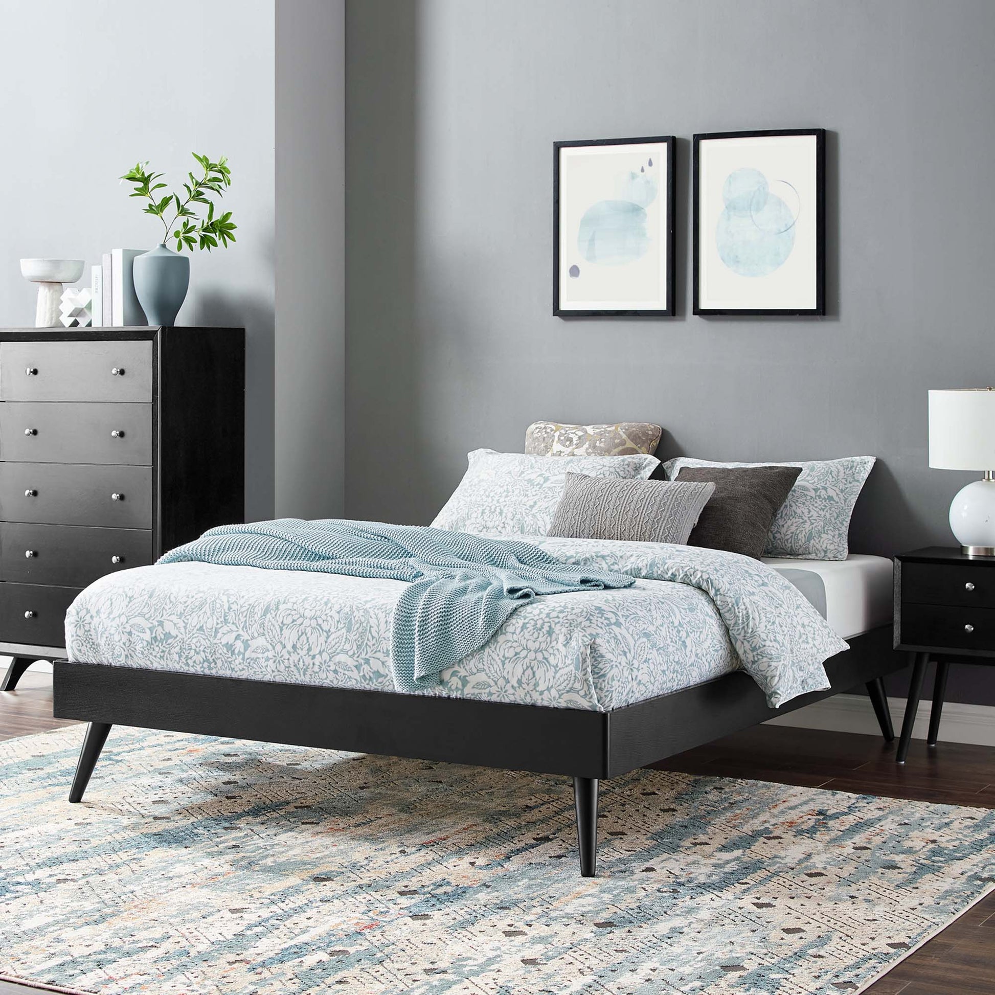 Low-profile black platform bed with tapered legs, blue floral bedding, pillows and a light blue throw in a modern bedroom.