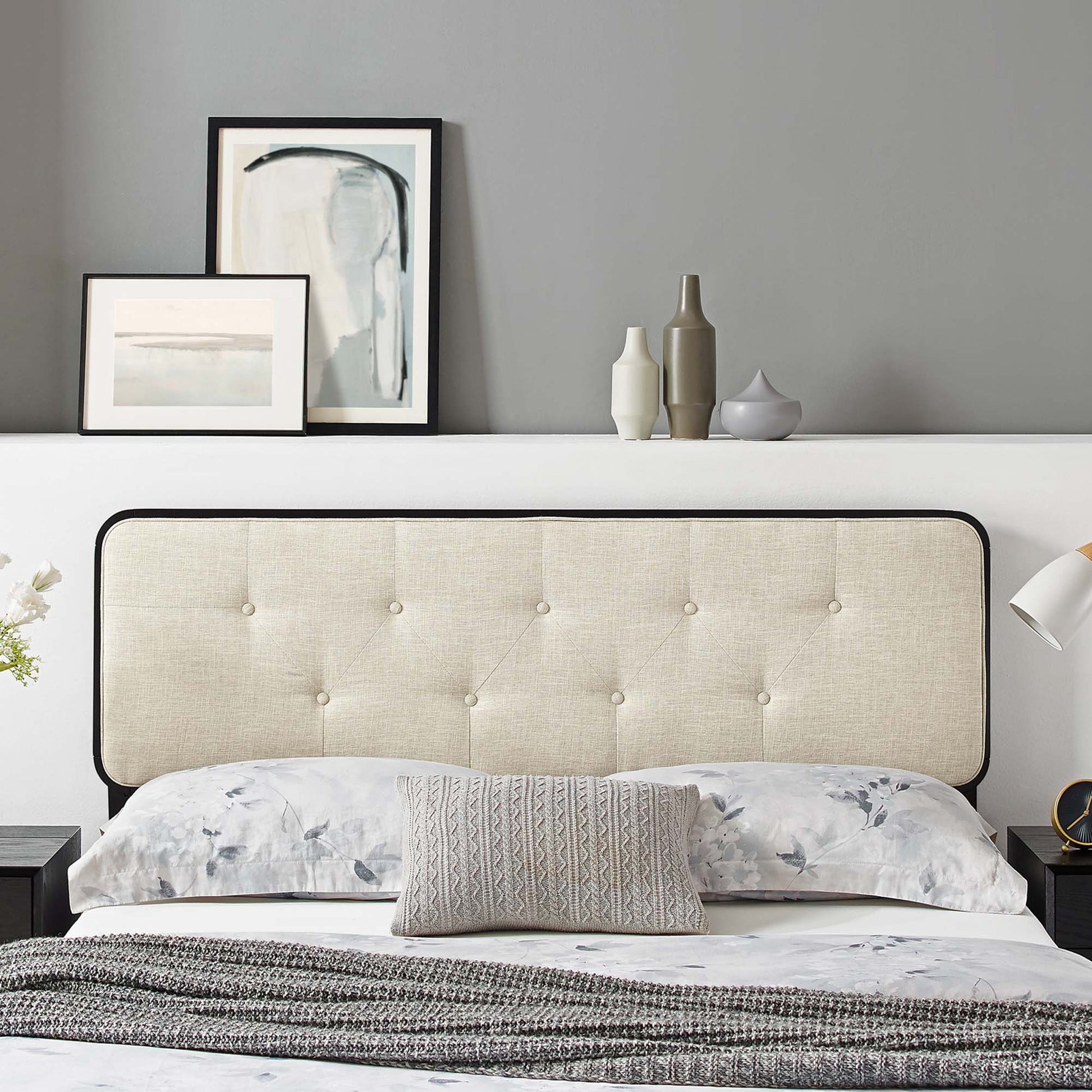Brown tufted headboard with black trim above a bed with floral bedding, gray knit throw, abstract art and vases on shelf.