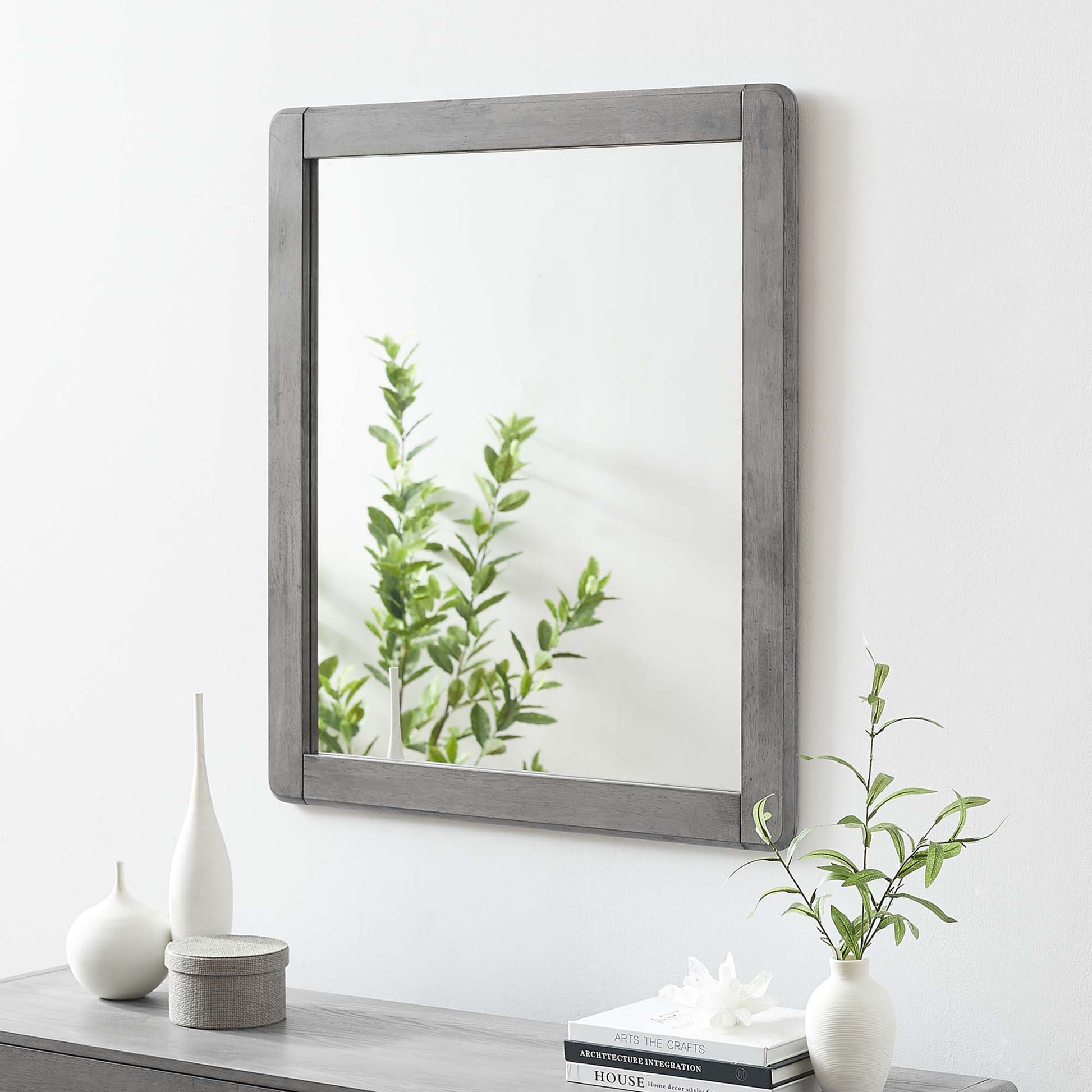 Rectangular rustic graywashed wood-framed mirror over a gray dresser with white vases, a small plant, and books.
