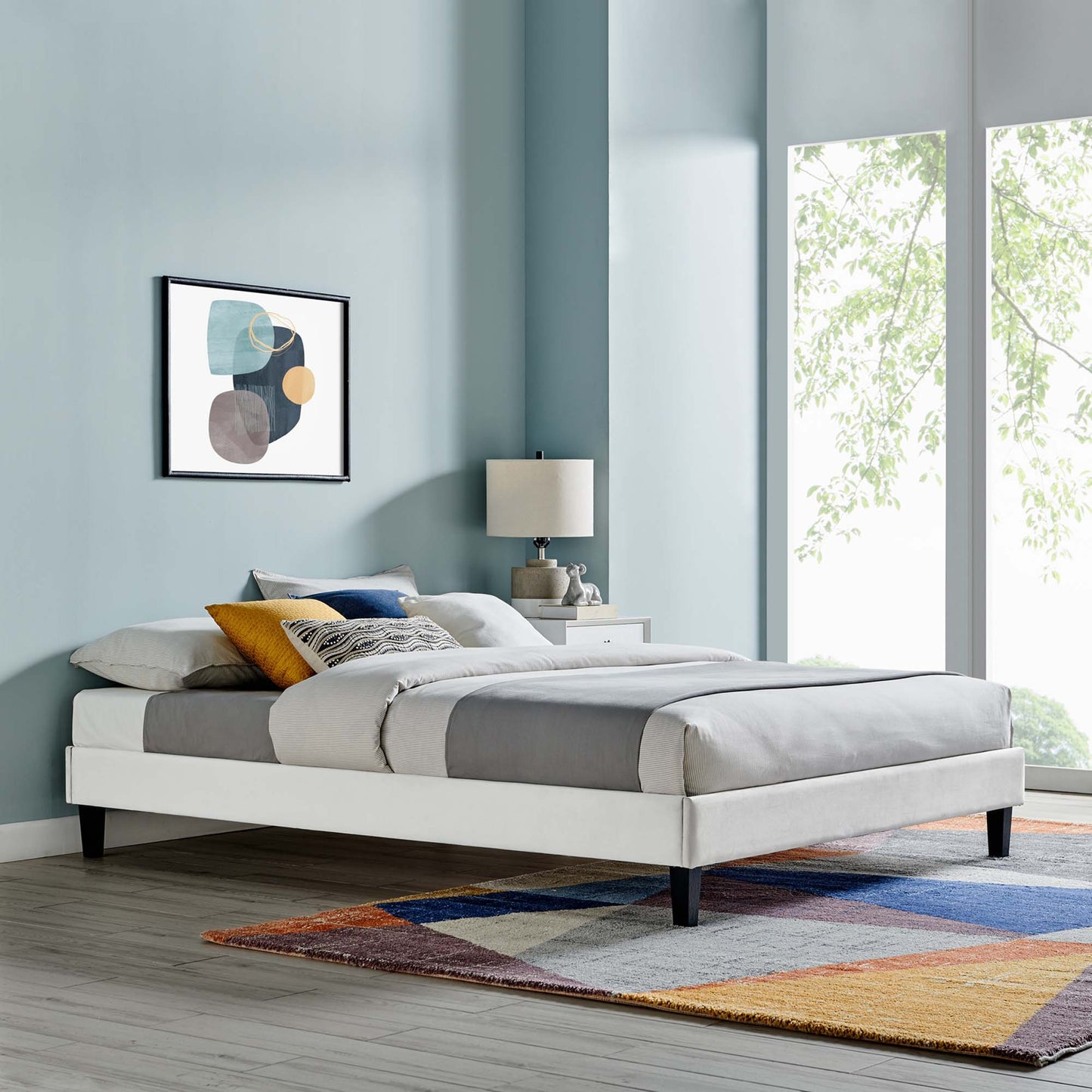 Light gray upholstered platform bed with dark legs, layered bedding, blue walls, colorful geometric rug, and abstract art.