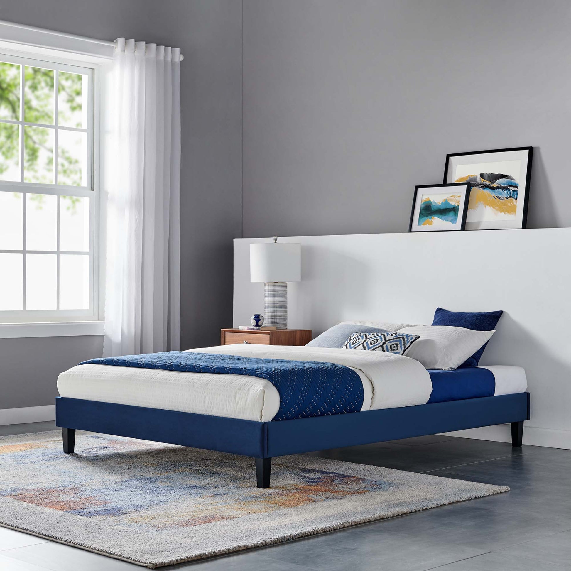 Navy upholstered full platform bed frame with black legs, white headboard, blue bedding, wooden nightstand, and abstract art.
