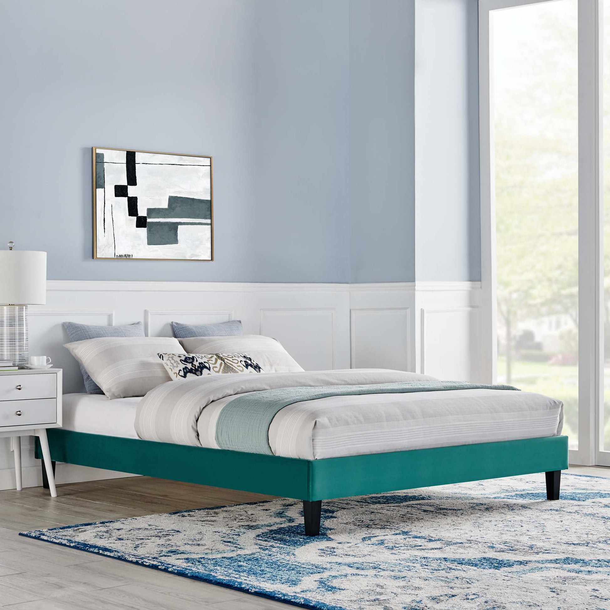 Teal upholstered platform bed with white and teal bedding in a blue-gray bedroom with abstract art and a blue rug.
