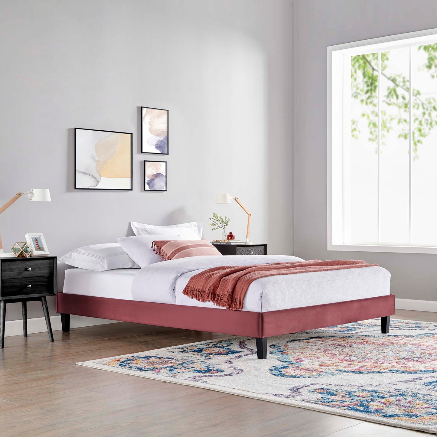 Maroon upholstered platform bed with white linens, pink throw, black nightstands, wooden arc lamps, abstract wall art, and colorful area rug