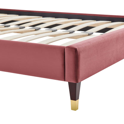Dusty rose velvet upholstered queen platform bed frame with dark wood legs and brass-tipped foot, slatted base visible.