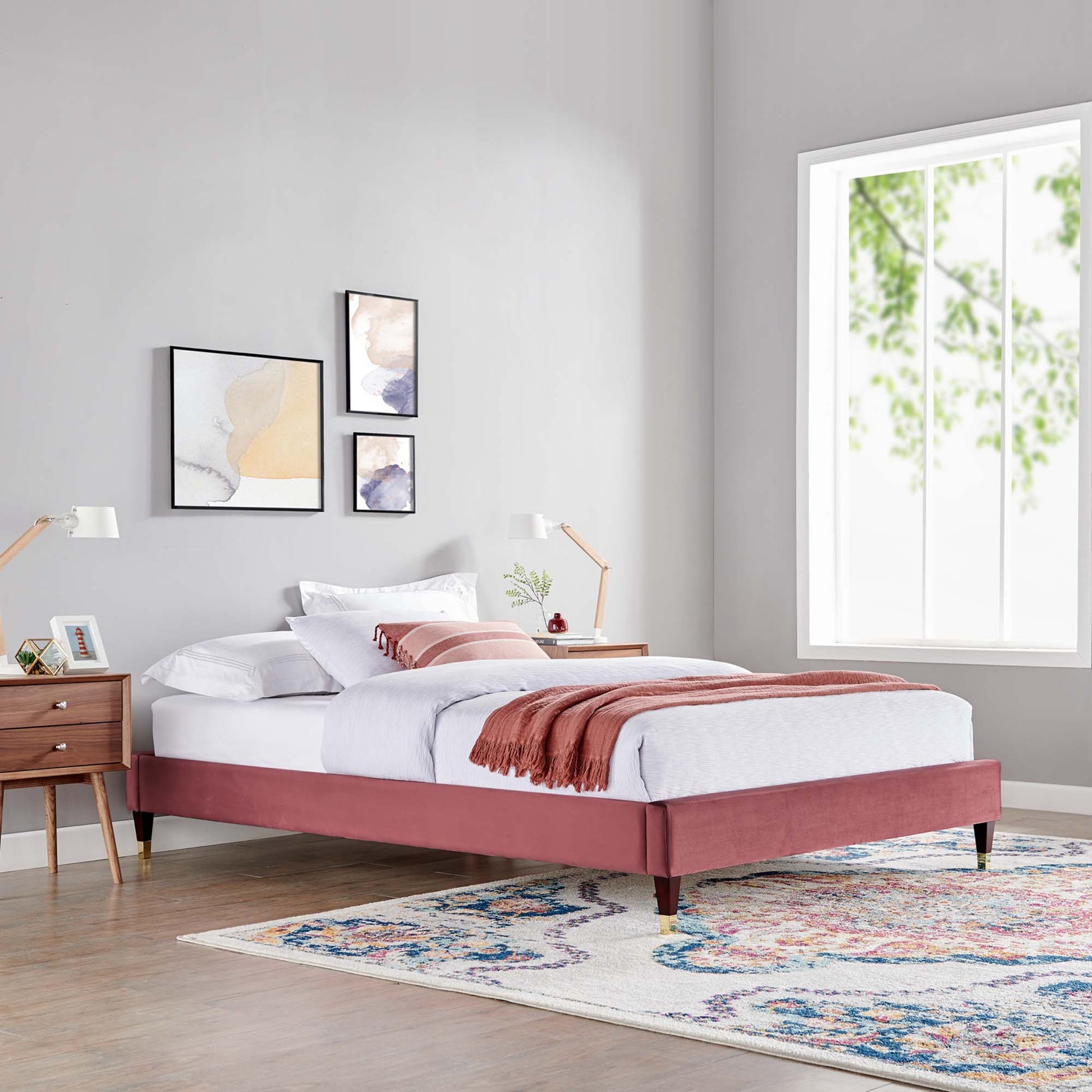 Dusty-rose upholstered platform bed with tapered legs, white linens, pink throw, in a gray modern bedroom with a colorful rug.