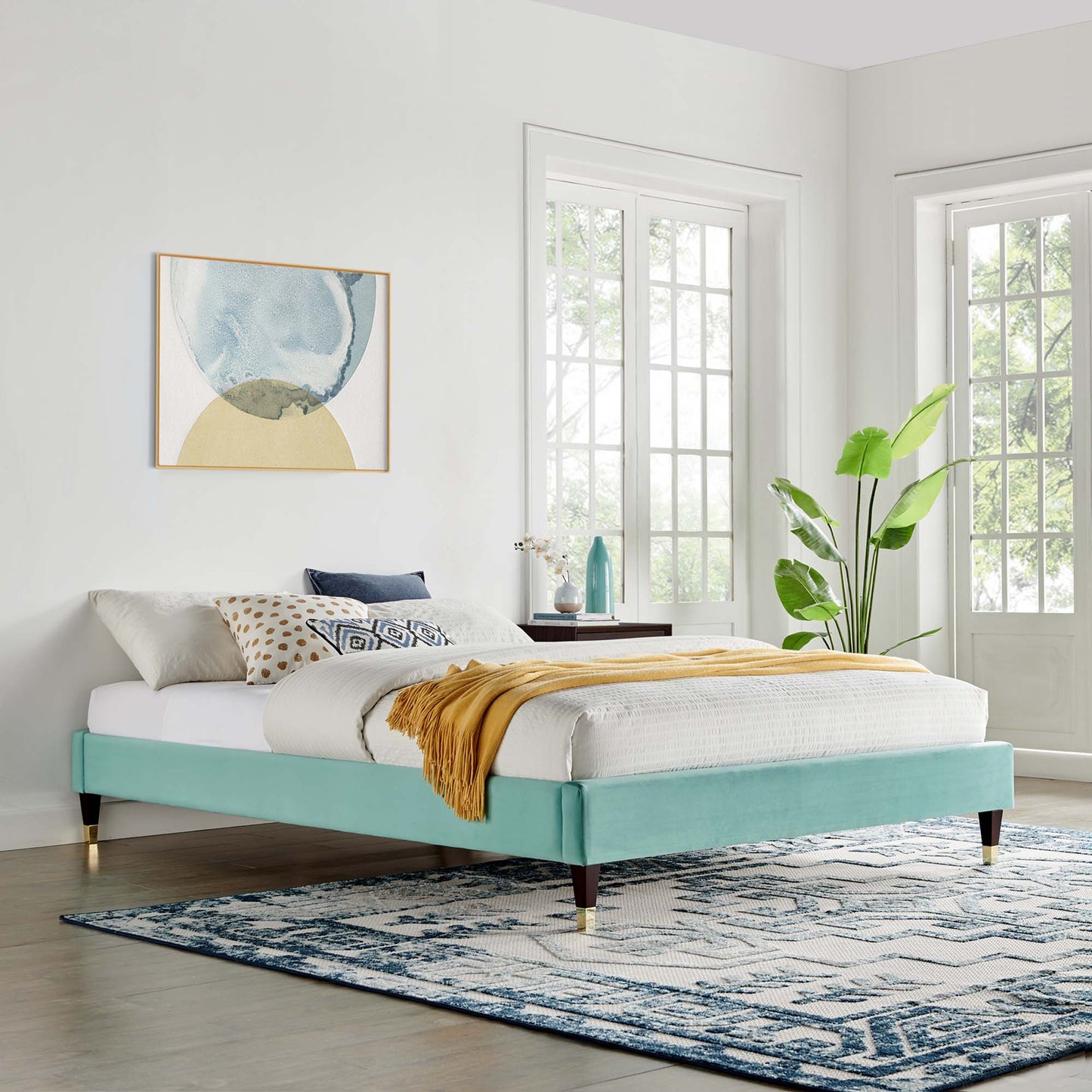 Mint velvet platform bed frame with white linens, mustard throw, geometric pillows, & brass-tipped legs in a bright bedroom.