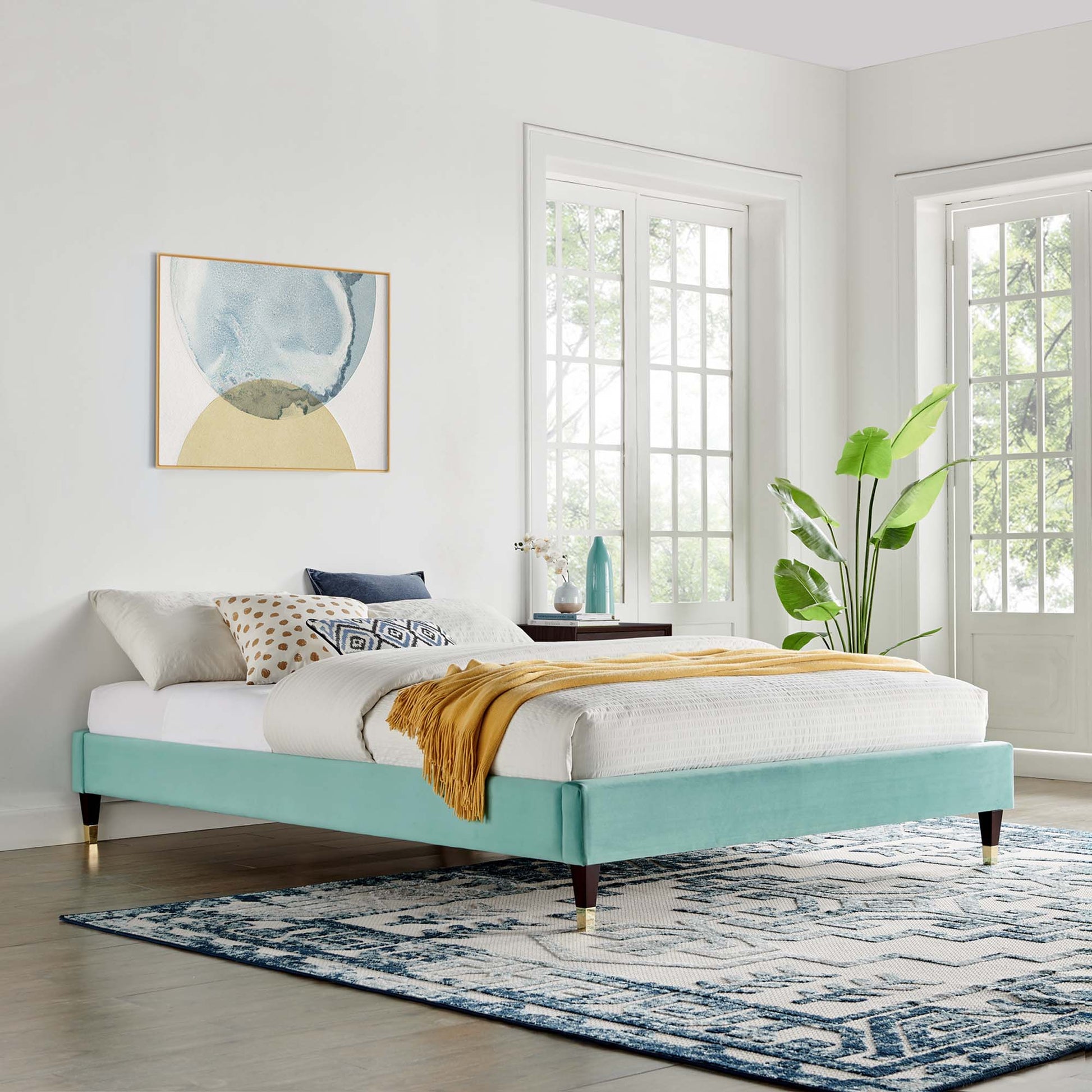Mint velvet platform bed frame with white linens, mustard throw, geometric pillows, & brass-tipped legs in a bright bedroom.