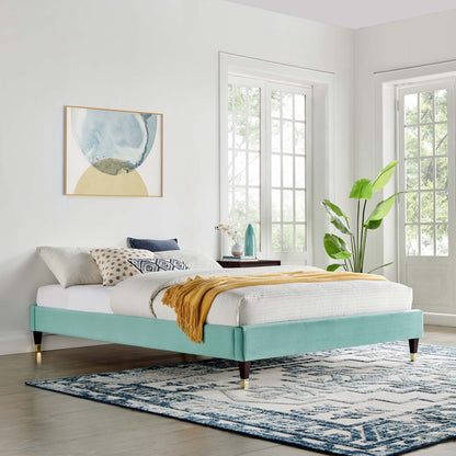 Mint velvet platform bed frame with white linens, mustard throw, geometric pillows, & brass-tipped legs in a bright bedroom.