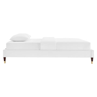 White upholstered platform bed with low-profile frame, wooden legs with brass tips, pillow and throw on bed.
