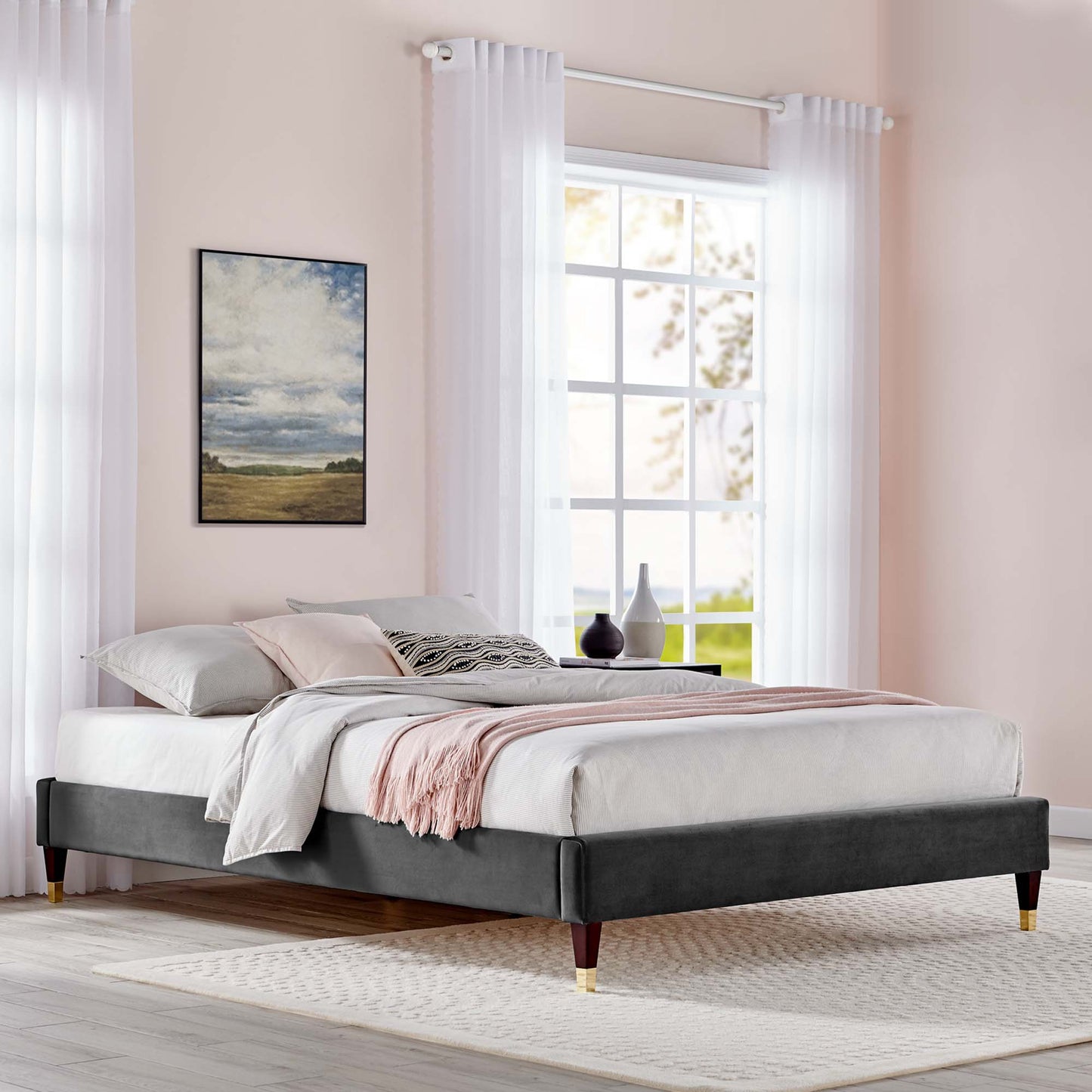 Charcoal velvet upholstered king platform bed frame with wooden tapered legs, white bedding, soft pink throw, in a pastel bedroom with sheer curtains.