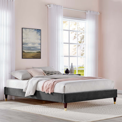 Charcoal velvet upholstered king platform bed frame with wooden tapered legs, white bedding, soft pink throw, in a pastel bedroom with sheer curtains.