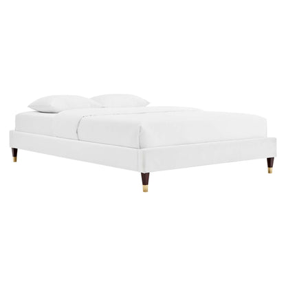 White upholstered platform bed with a low-profile frame, tapered wooden legs with brass caps, and white bedding with two pillows.