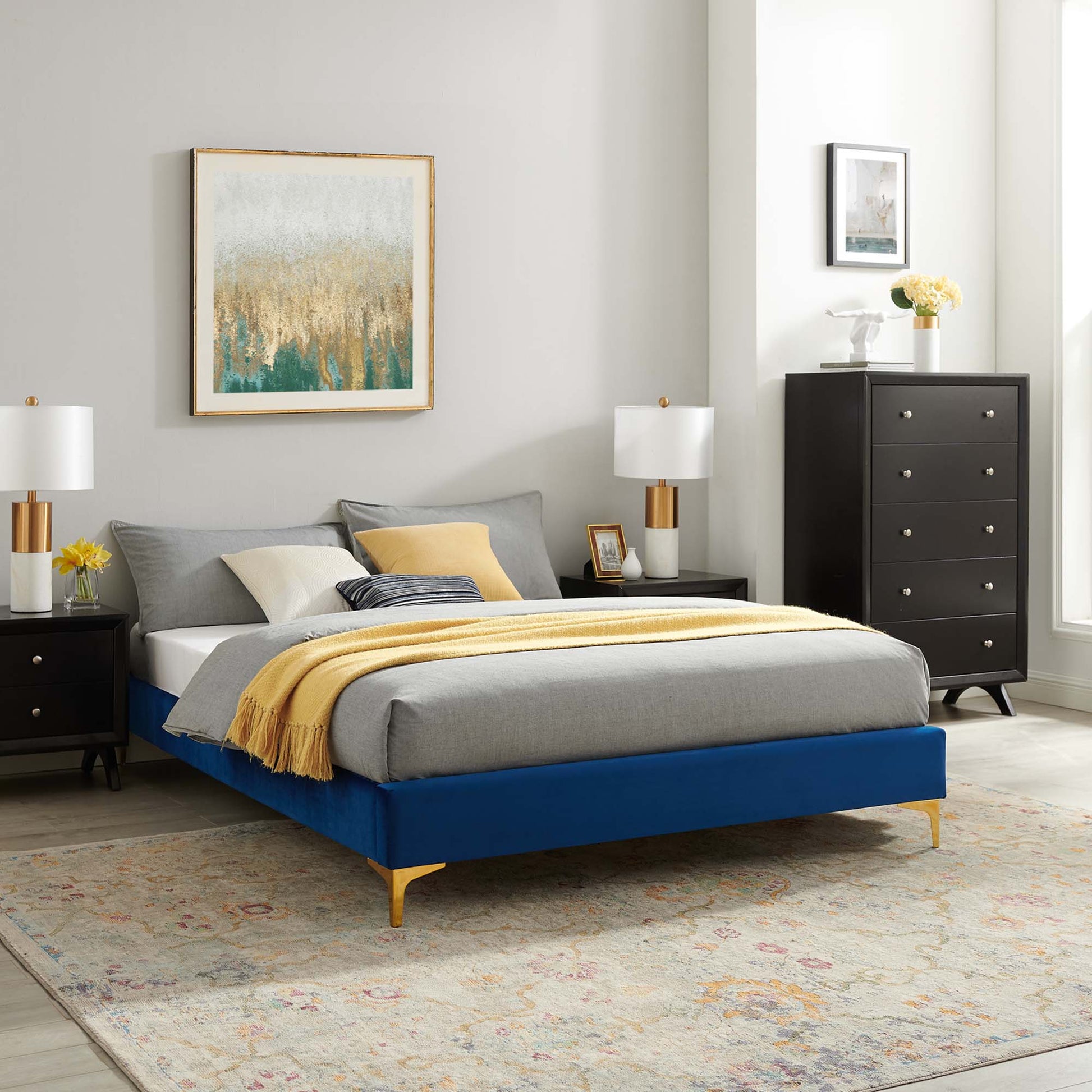 Navy upholstered platform bed with gold legs, gray bedding and a yellow throw in a modern bedroom.