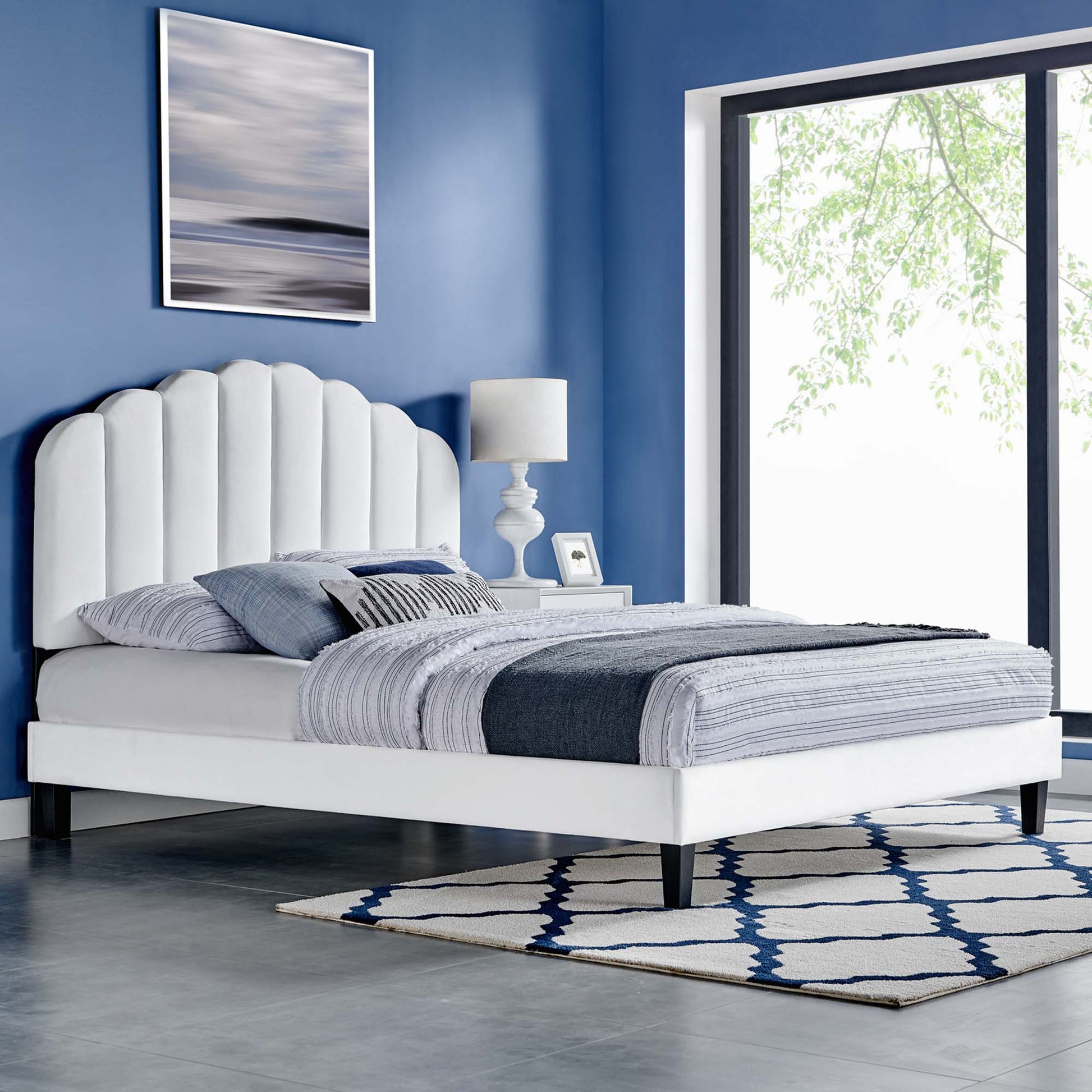 White upholstered queen platform bed with scalloped headboard in blue walls, gray-blue bedding, nightstand lamp, geometric rug.