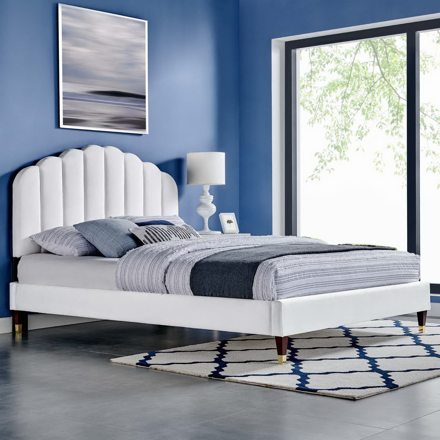 White upholstered platform bed with scalloped headboard, blue bedding, nightstand lamp, and a geometric rug.