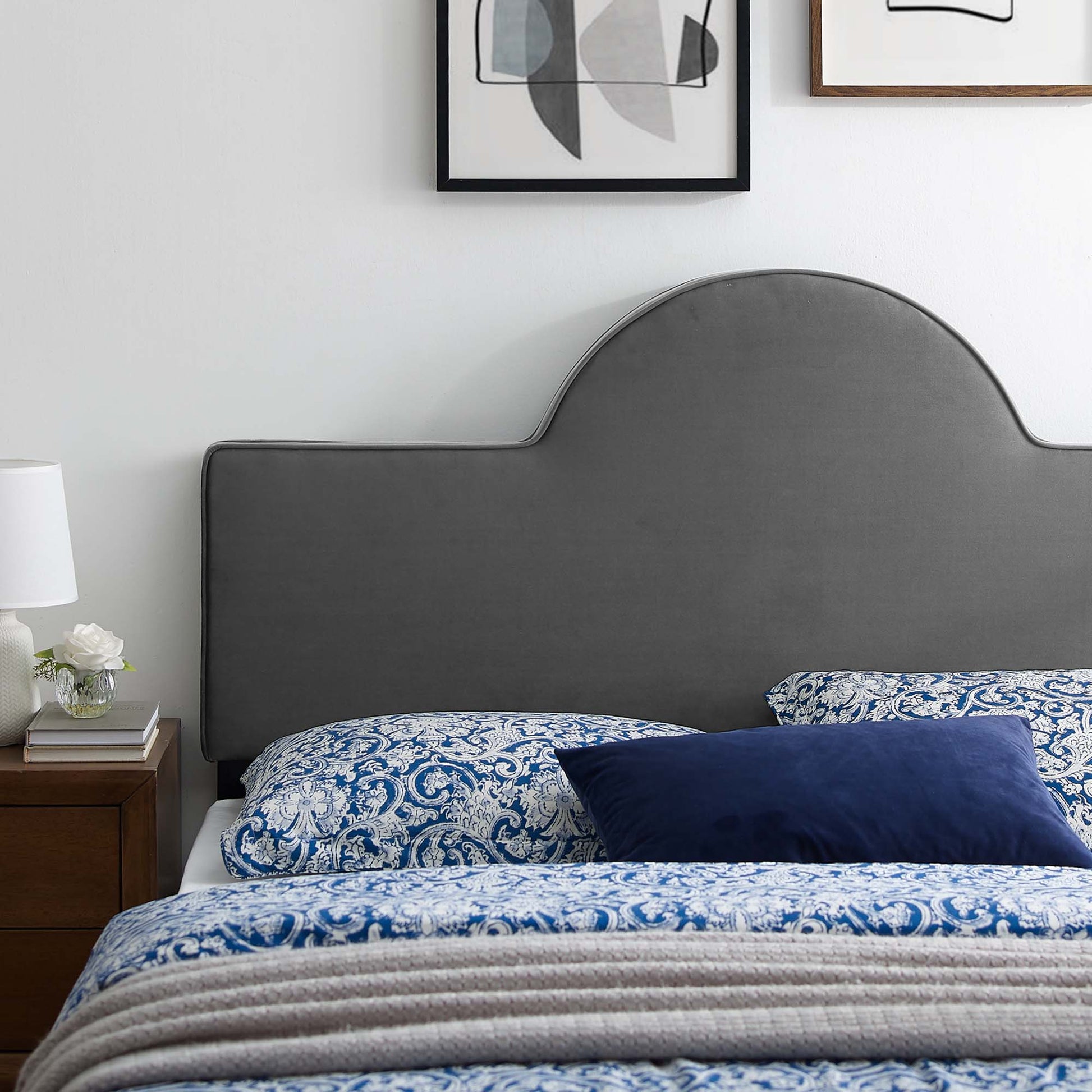 Grey curved upholstered twin headboard with blue paisley bedding, navy throw pillow, wooden nightstand, lamp and framed art.