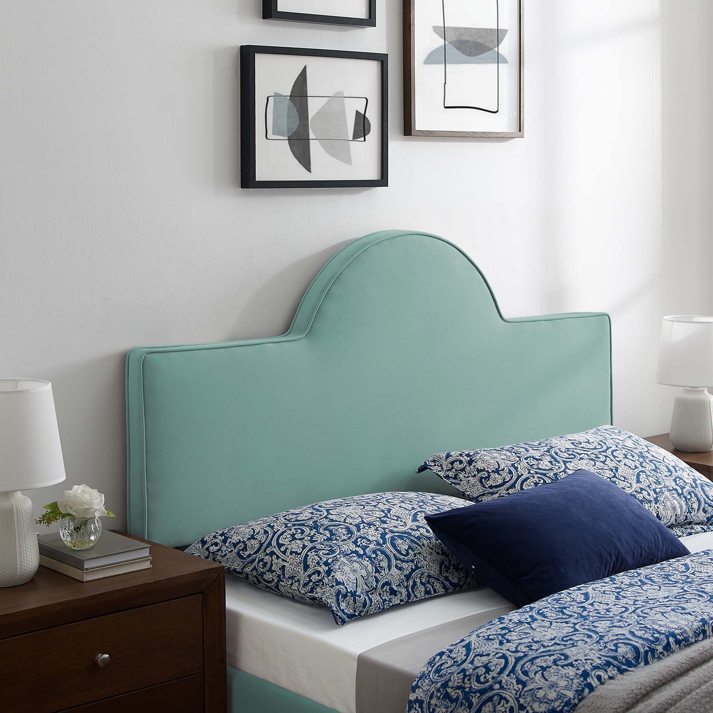 Mint green upholstered headboard with curved arch, blue paisley bedding, dark wood nightstand, white lamps, abstract art.
