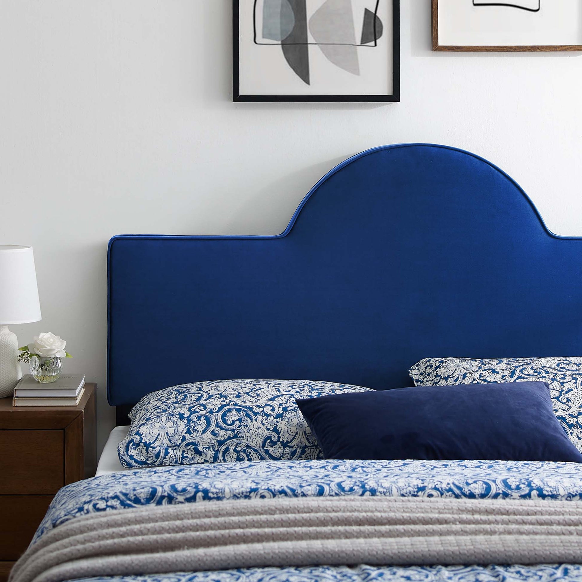 Navy curved upholstered headboard with blue damask bedding, beige throw, wooden nightstand, lamp, and framed art.