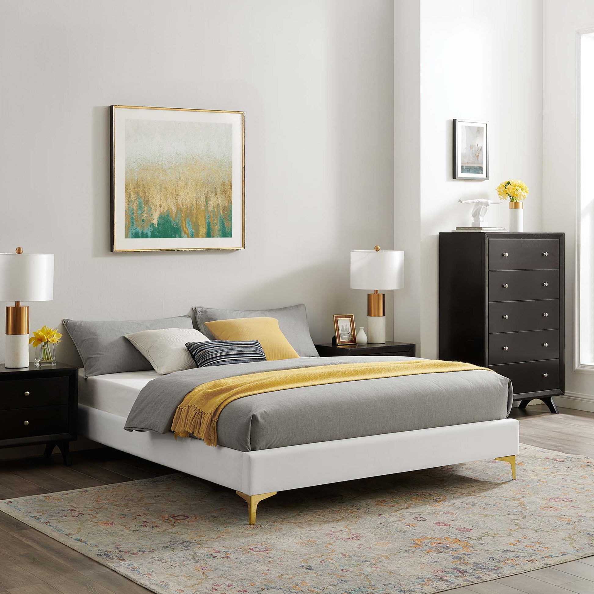 White platform bed with gray bedding, yellow throw, black nightstands, brass lamps, tall dresser, abstract wall art, rug