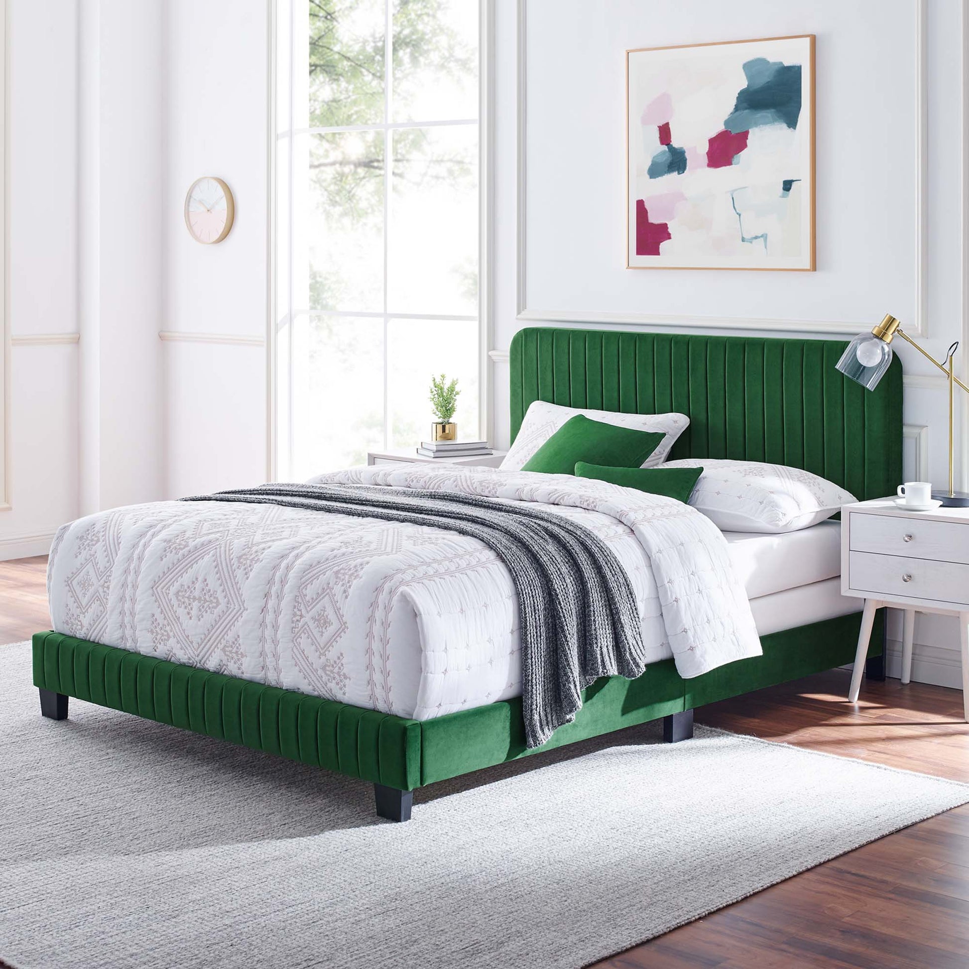 Emerald green velvet bed with tufted headboard, white patterned duvet, gray throw, white nightstands, gold lamp, abstract art.