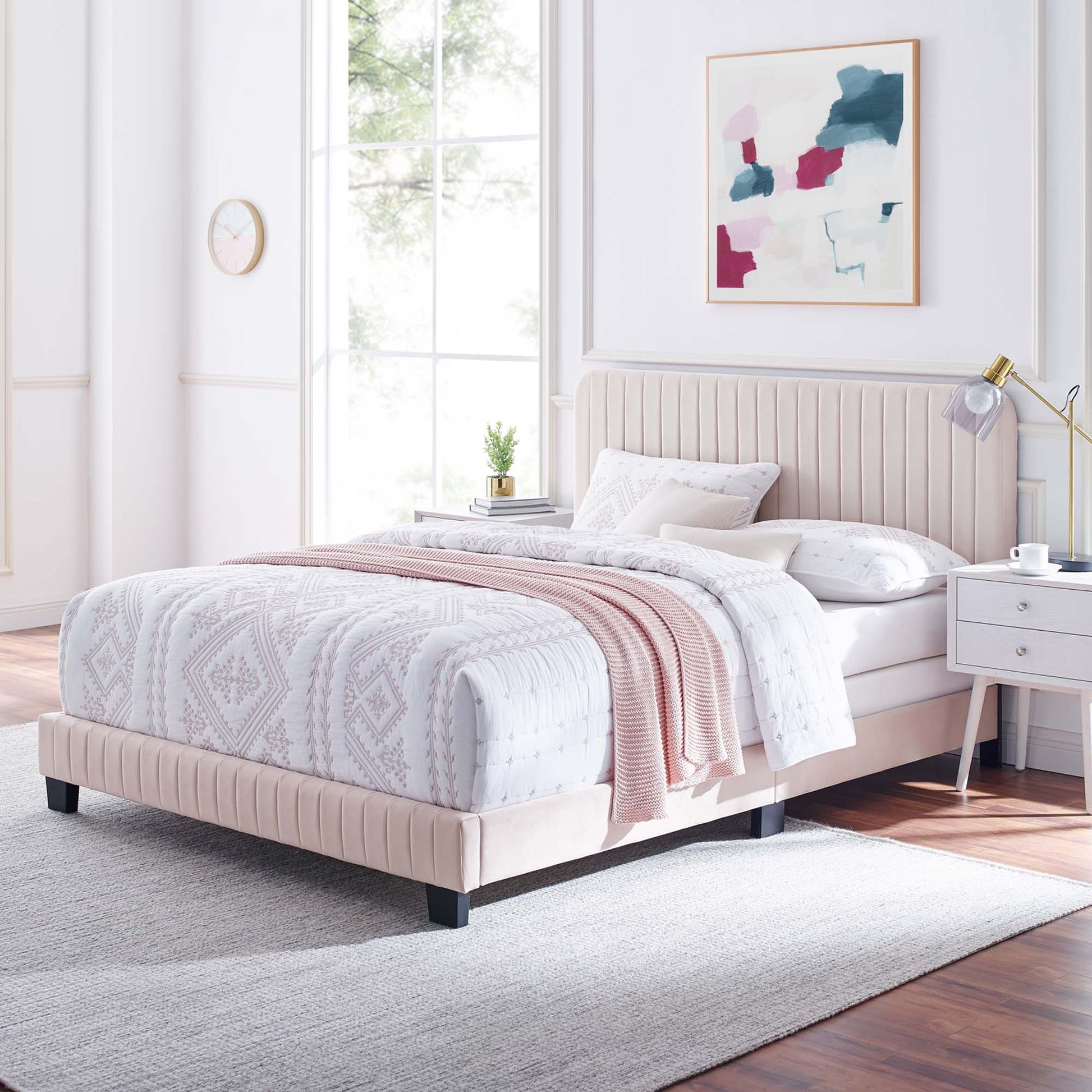 Pastel pink upholstered platform bed with tufted headboard, white geometric bedding, blush knit throw, modern bedroom.