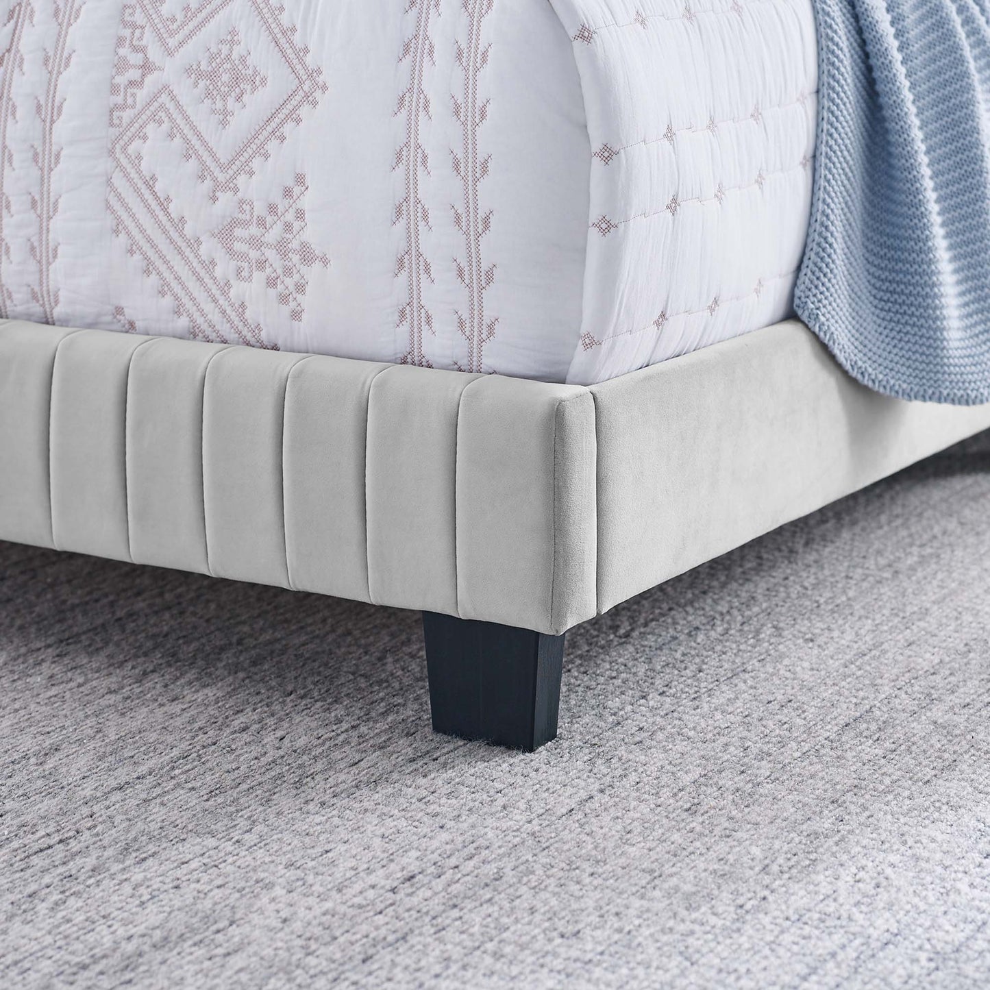 Light gray upholstered full bed with vertical channel tufting and dark legs, dressed with a white quilt with pink embroidery and a blue throw.