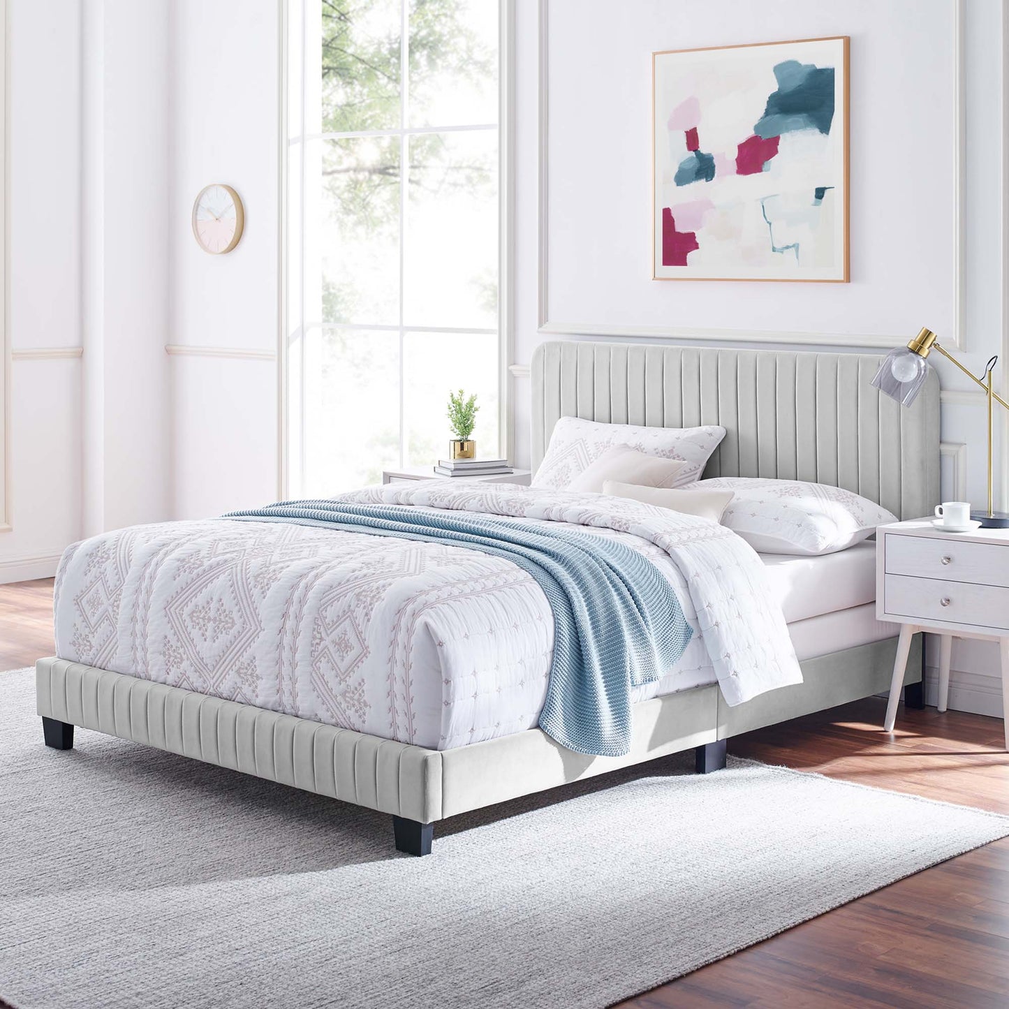 Light gray upholstered full bed with tufted headboard in a bright modern bedroom, white-pink bedding and a blue throw.