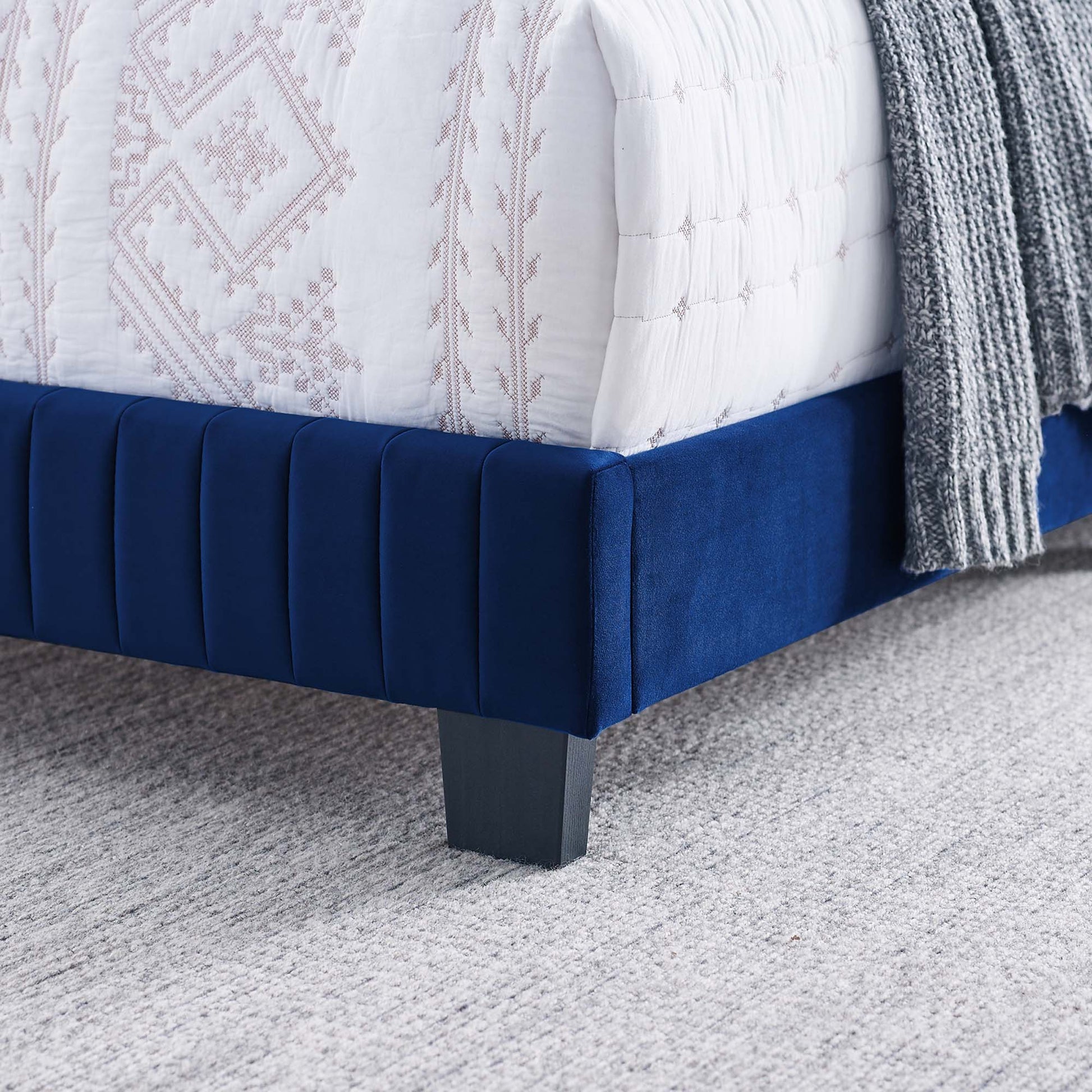 Navy blue velvet upholstered bed base with vertical channel tufting and a dark square leg; white quilt and gray knit throw.
