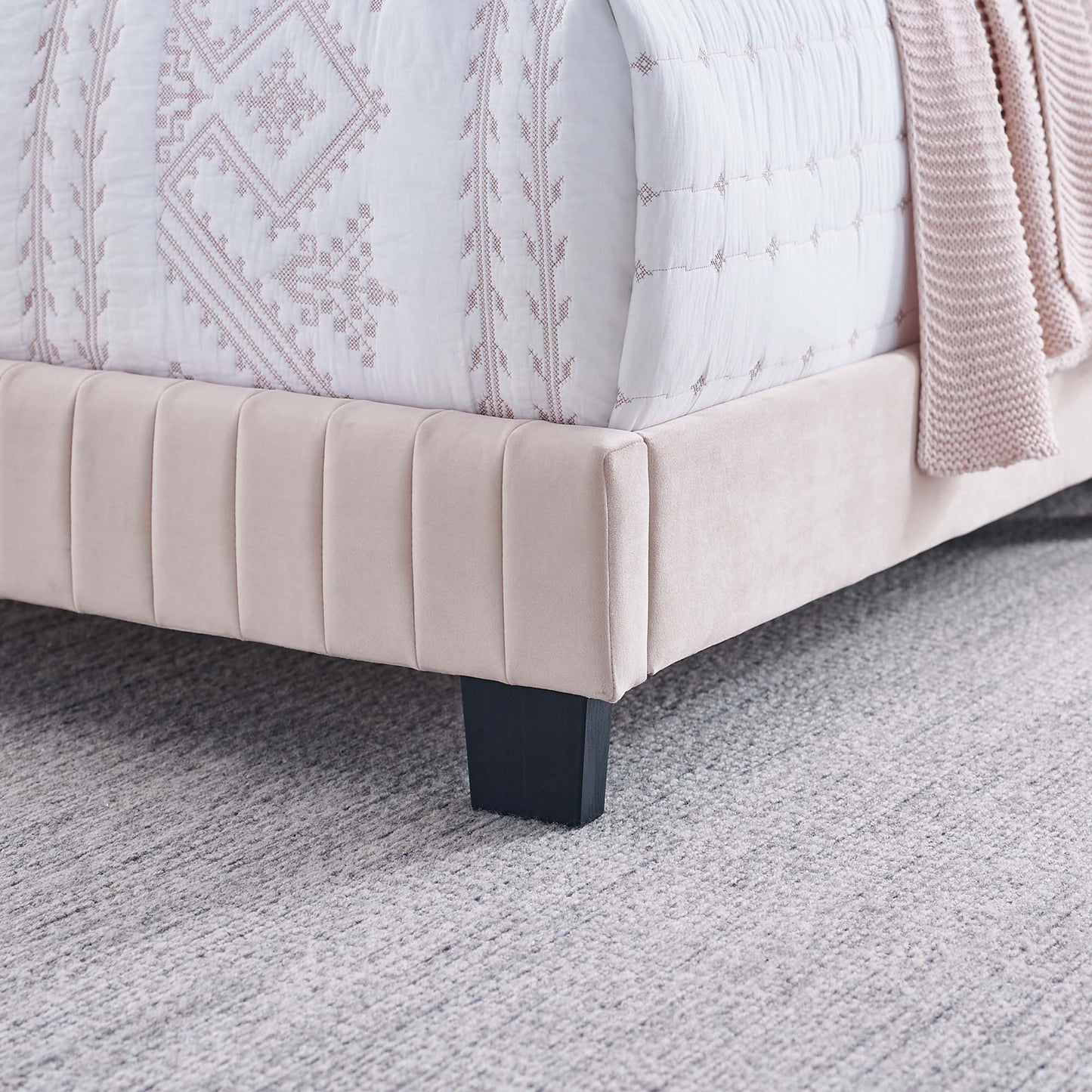 Blush pink upholstered bed base with vertical panels, dark block leg, white quilt with pink embroidery, soft pink throw