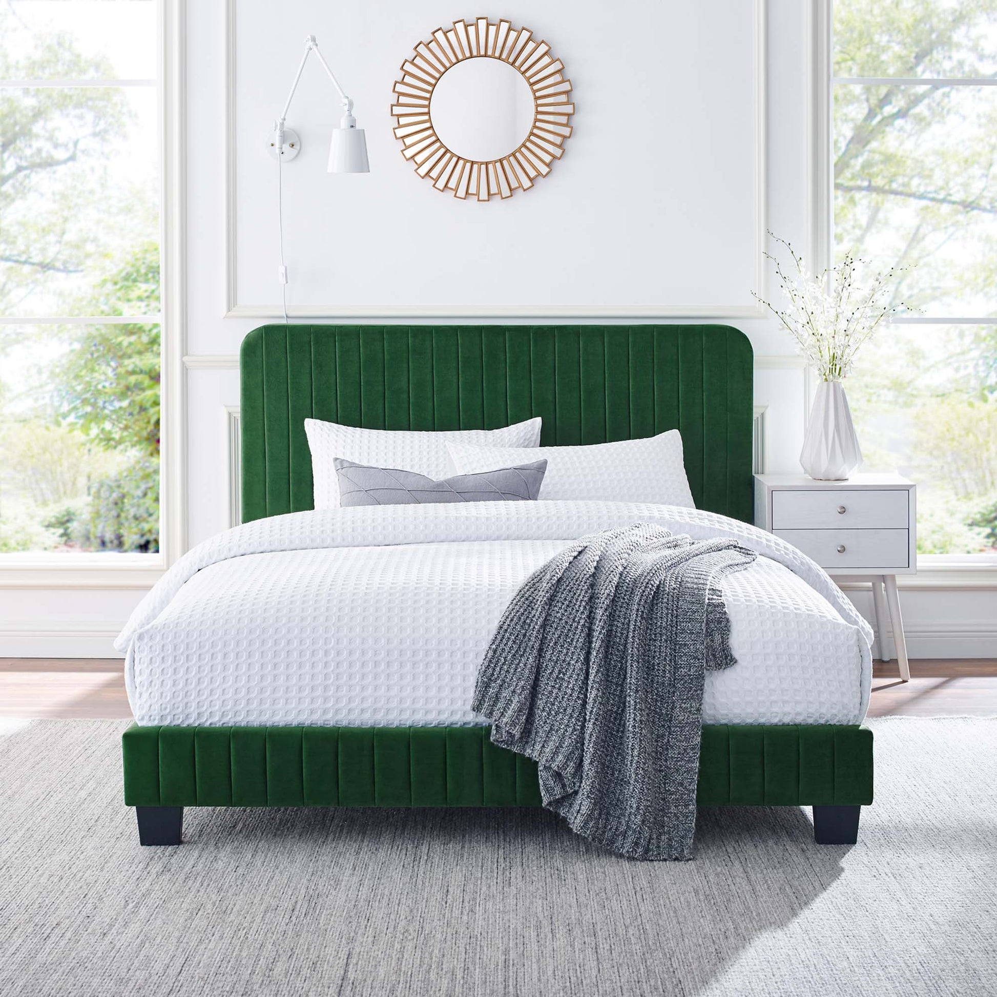 Emerald green velvet bed with tufted headboard, white textured bedding, gray knit throw, sunburst mirror above.