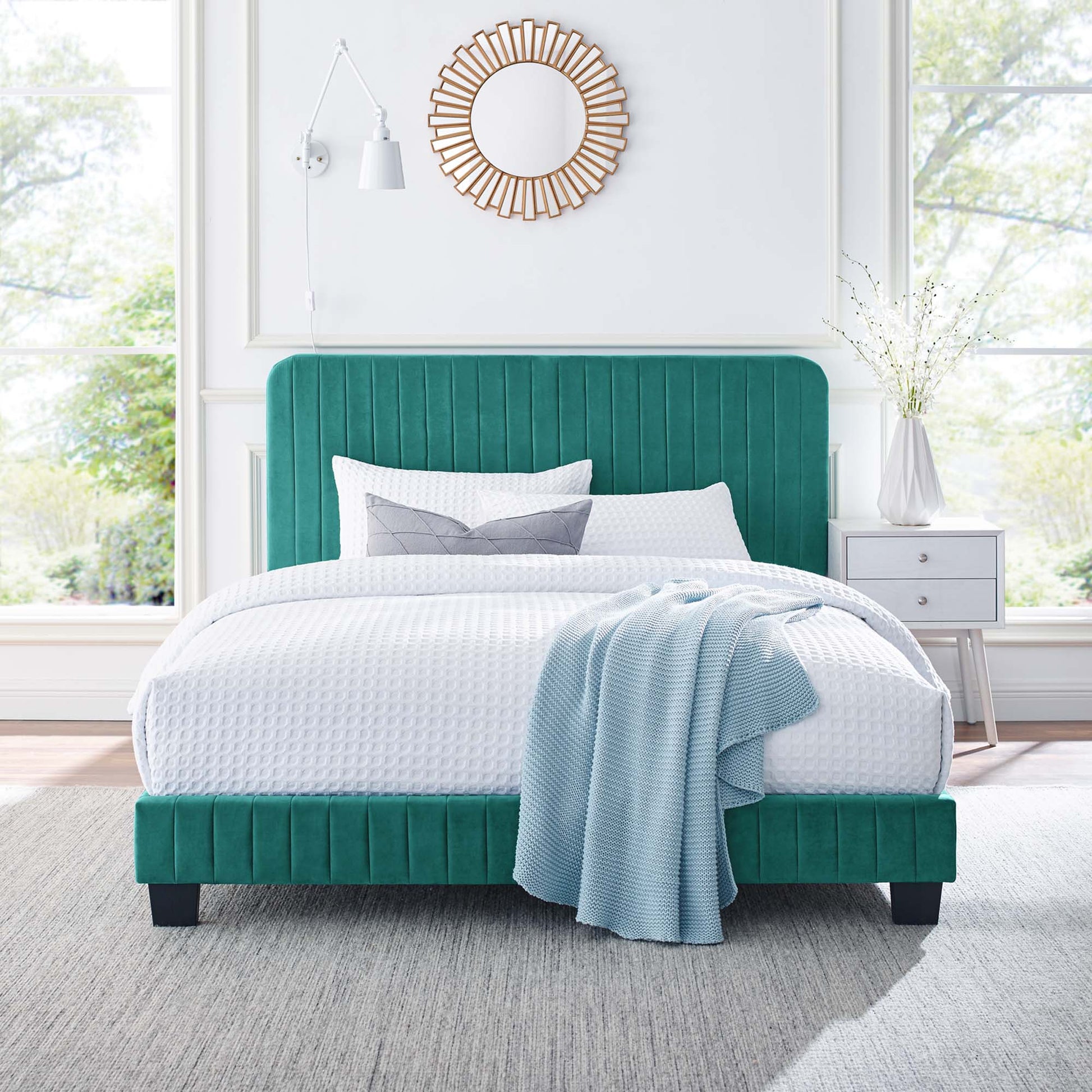Teal upholstered bed with vertical-channel headboard, white bedding, light blue throw, round sunburst mirror above.