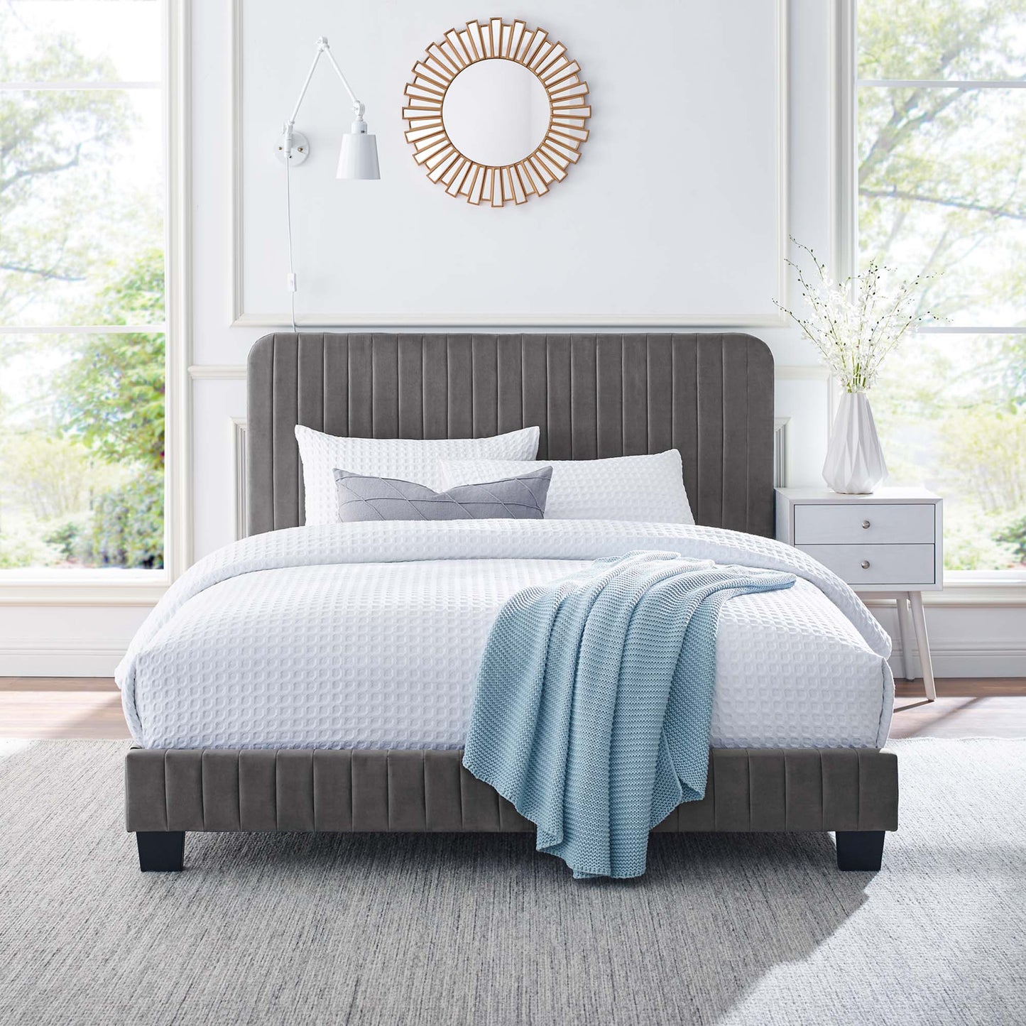 Gray upholstered platform bed with tall tufted headboard, white bedding, light blue knit throw, and sunburst mirror above.