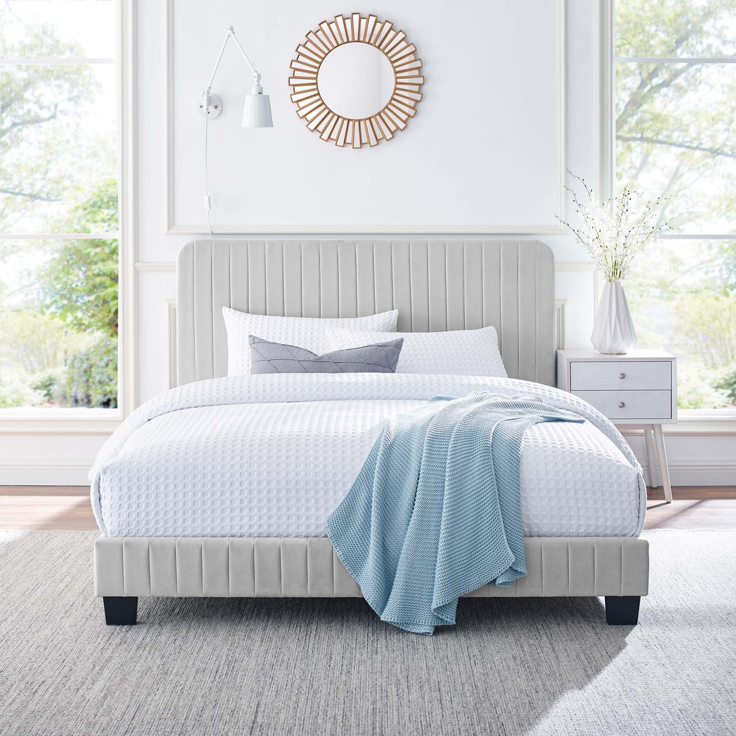 Light gray upholstered platform bed with vertical tufting, white bedding, blue throw, and sunburst mirror in an airy room.