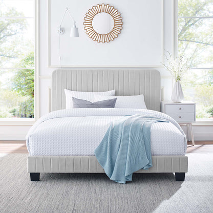 Light gray upholstered platform bed with vertical tufting, white bedding, blue throw, and sunburst mirror in an airy room.