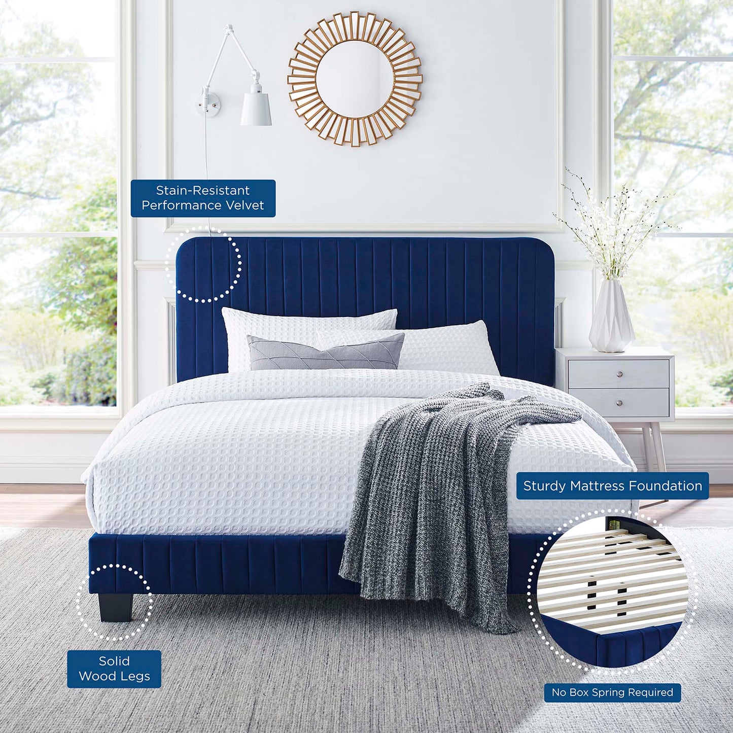 Navy velvet upholstered platform bed with solid wood legs, white bedding and gray throw; sunburst mirror overhead.