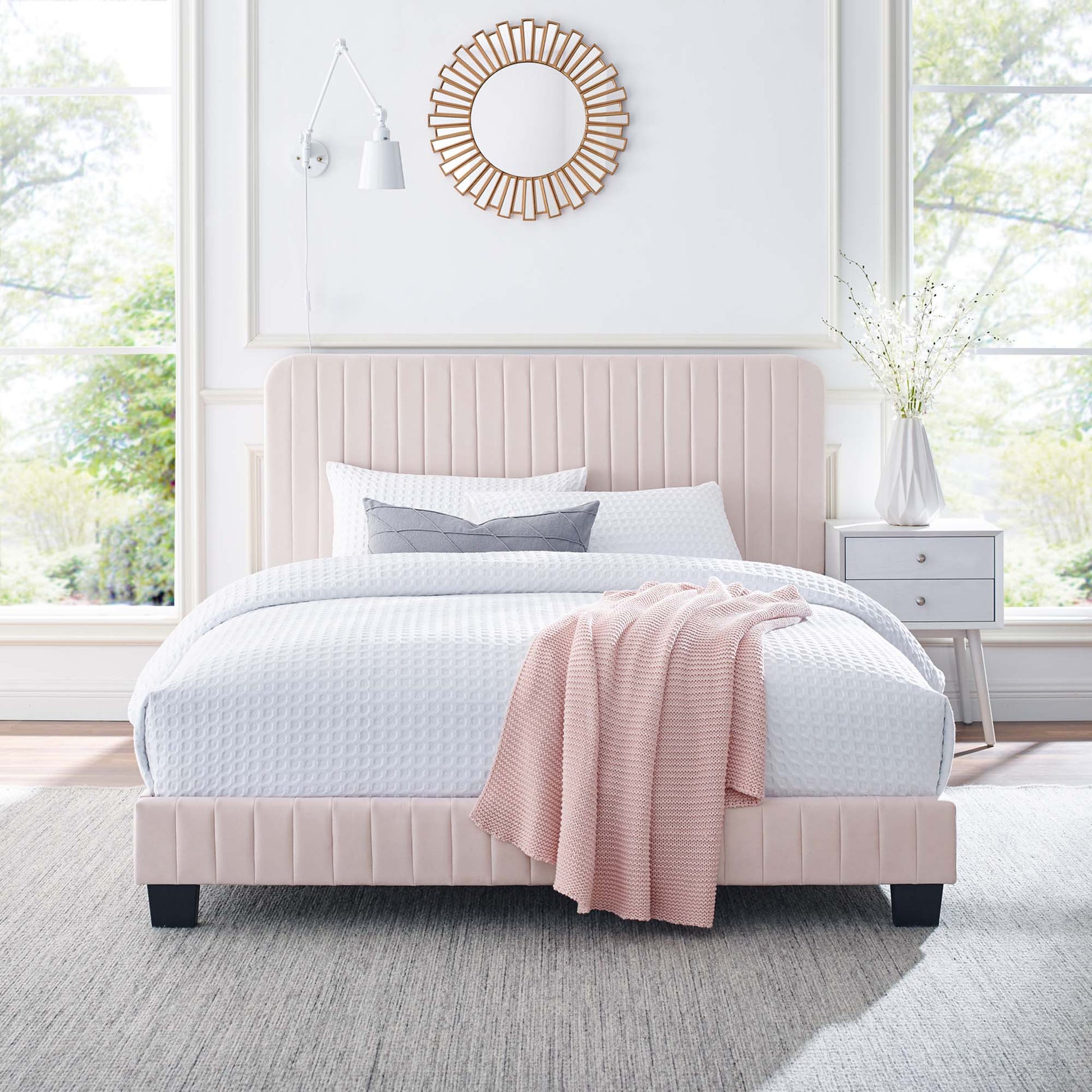 Pink upholstered full-size platform bed with a tall padded headboard, white quilted bedding, grey pillows, pink knit throw, and dark legs.