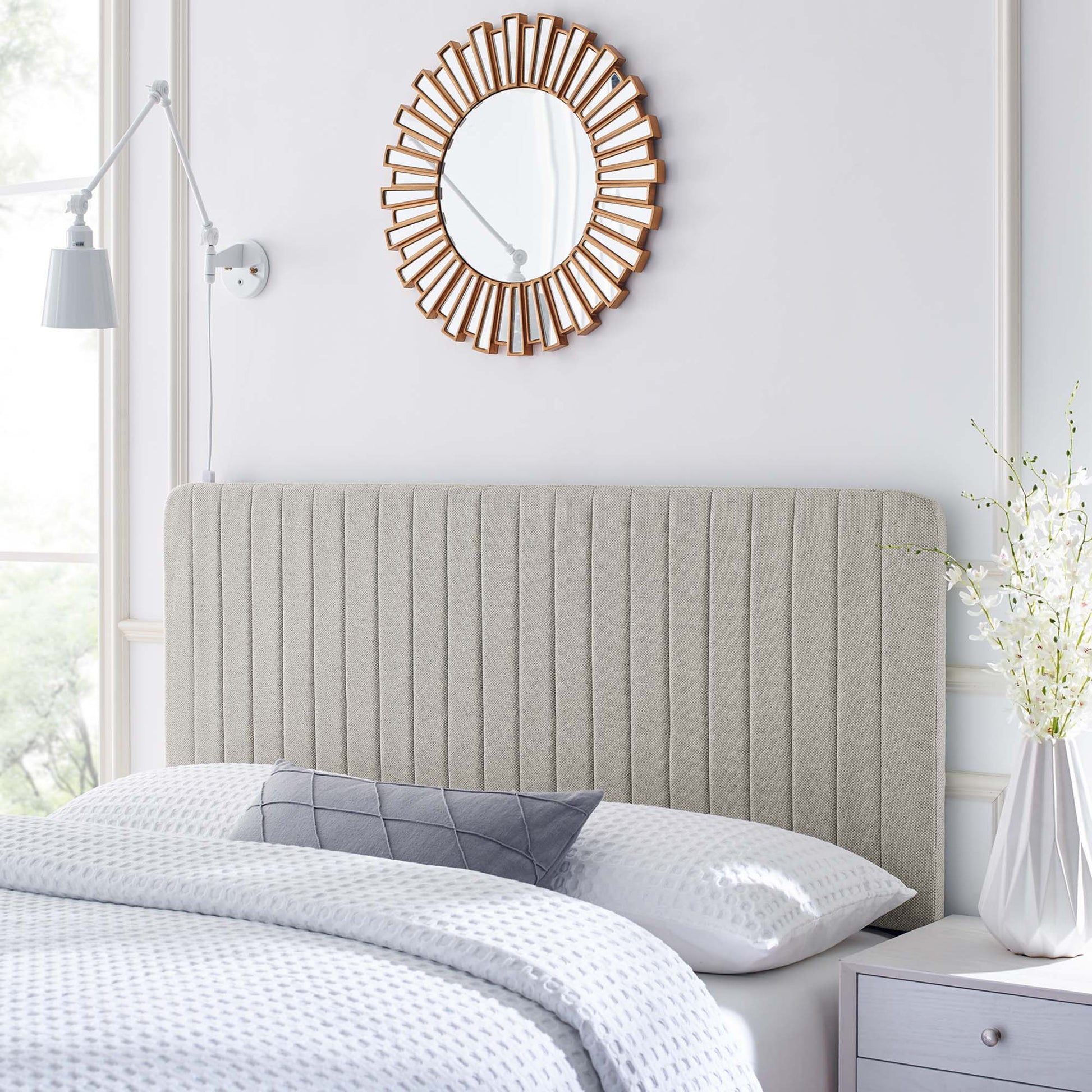 Grey upholstered vertical-channel headboard with white bedding, sunburst wood mirror, white nightstand, vase of flowers.
