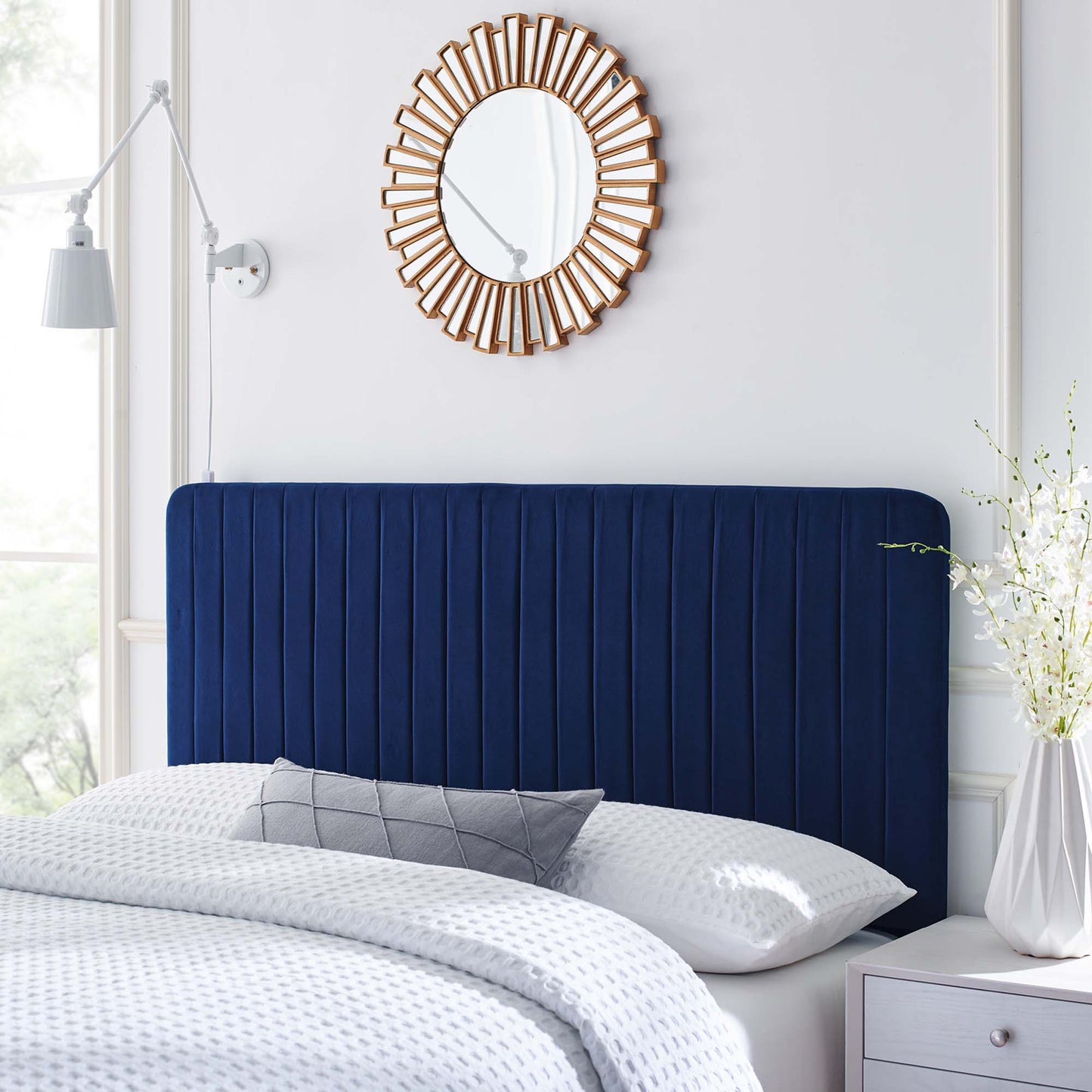 Navy velvet headboard with vertical channels, white textured bedding, gray pillow, sunburst wooden mirror, white nightstand.