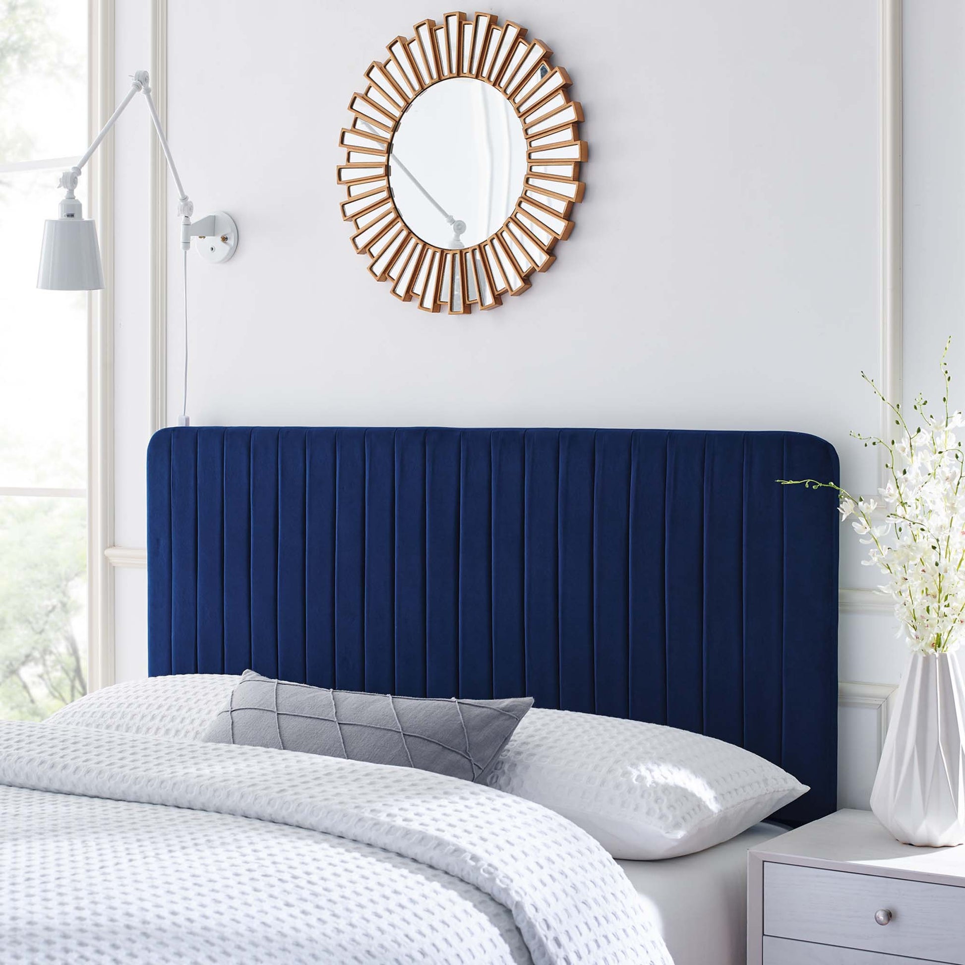 Navy velvet headboard with vertical channels, white textured bedding, gray pillow, sunburst wooden mirror, white nightstand.