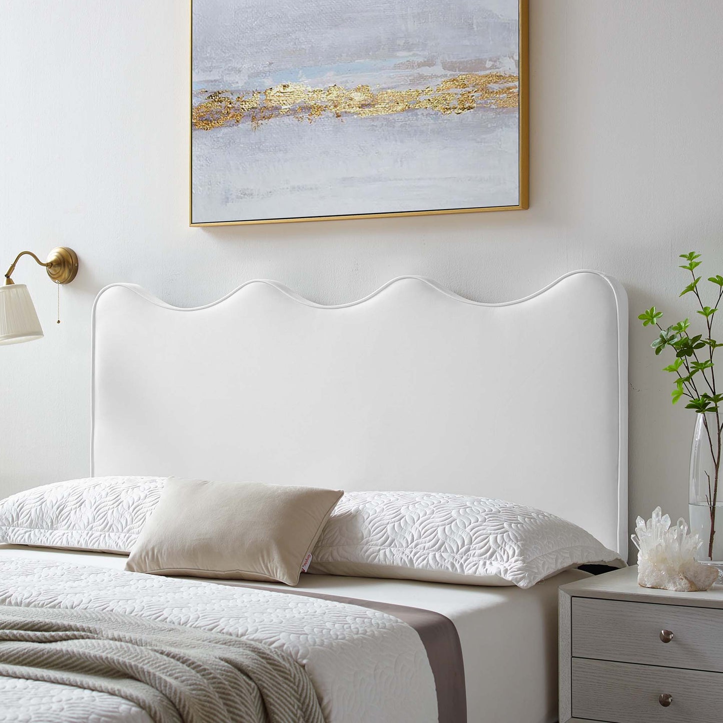 White scalloped headboard with quilted bedding, beige pillow, taupe throw, brass lamp, abstract art, gray nightstand.