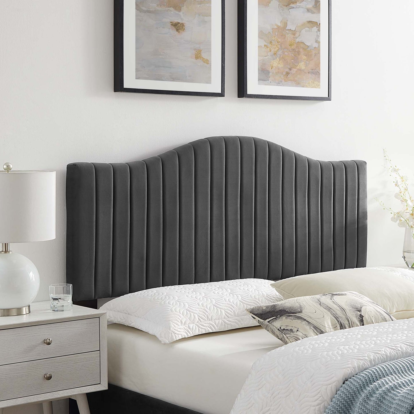 Dark gray vertical-channel upholstered headboard with curved top, beside a light gray nightstand and white quilted bedding.